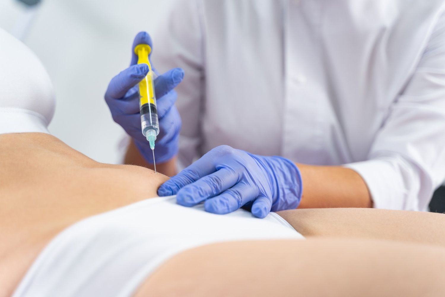 Doctor administering a medical injection near a patient’s waist, wearing blue gloves and holding a syringe.