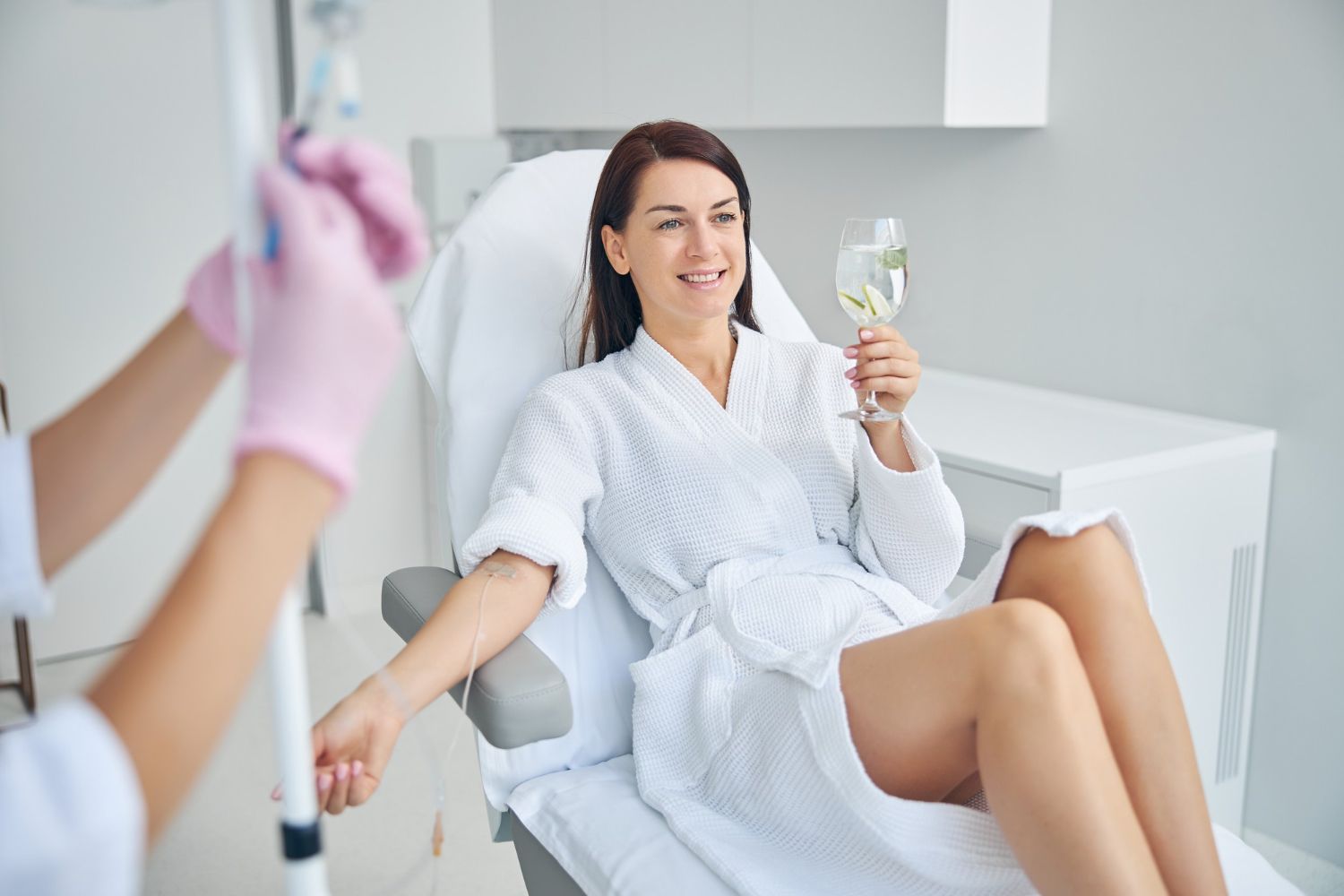 Woman in white robe relaxing in a clinic chair while holding a glass and smiling, with a nurse nearby
