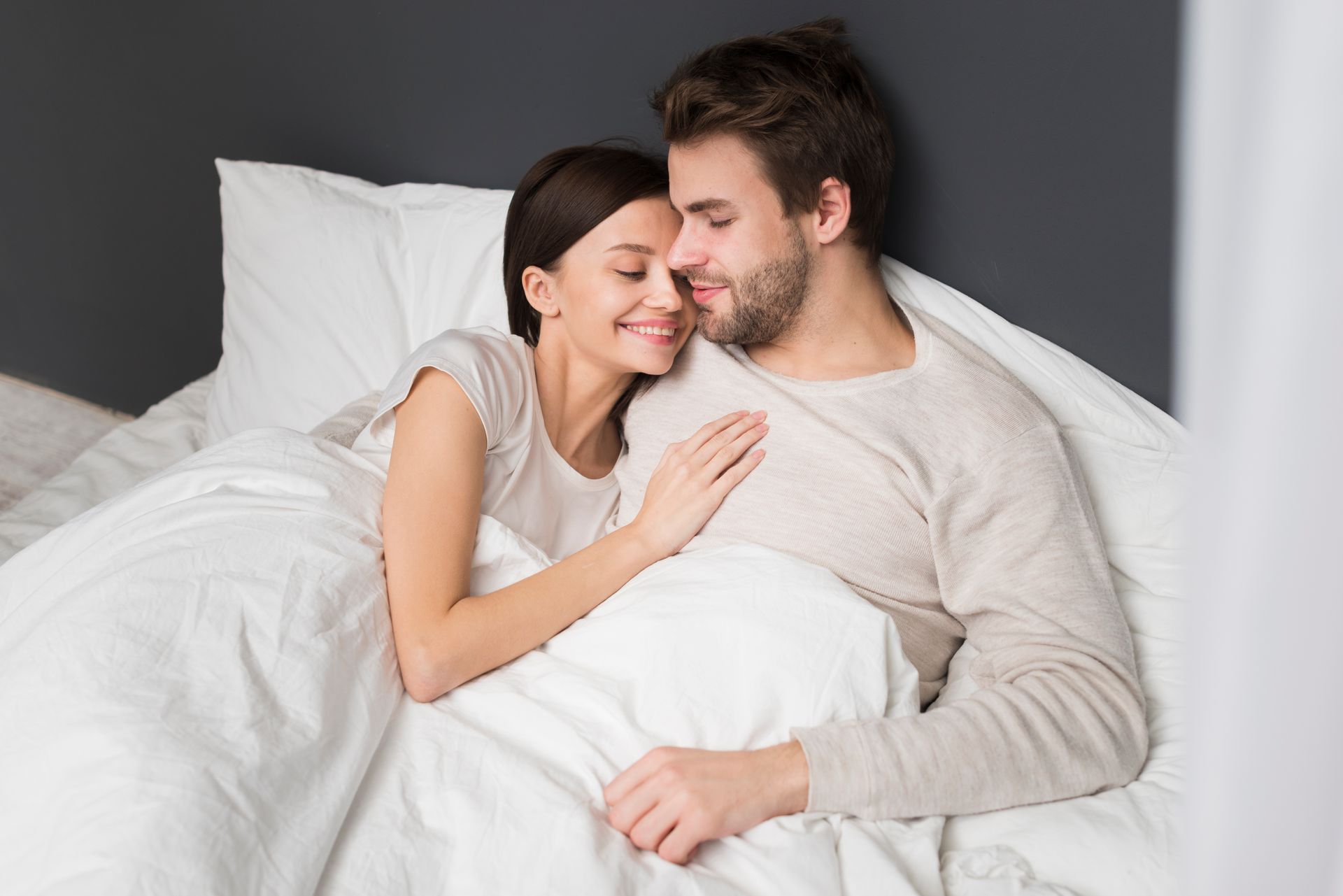 Couple cuddling in bed; woman smiles, rests head on man's chest, both under white covers.