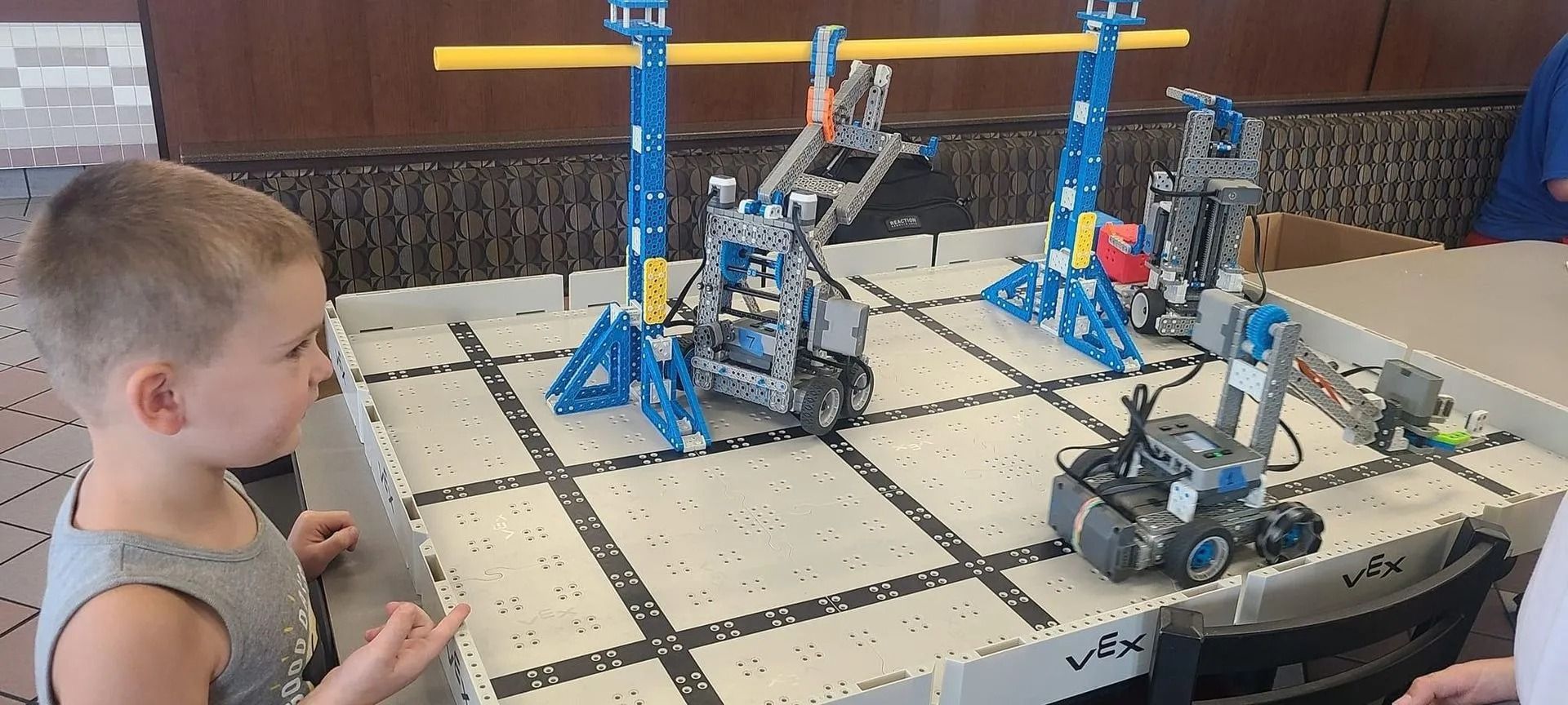 Boy watches robotic contraptions on a competition table. Blue and grey robots, yellow beam.