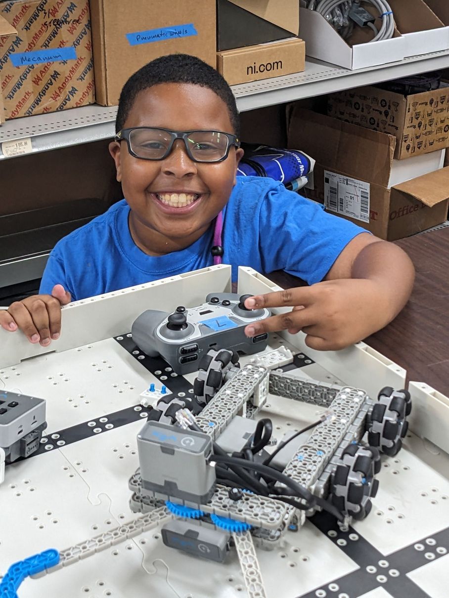 Boy with glasses smiling, operating a robot with a remote control, in a classroom setting.