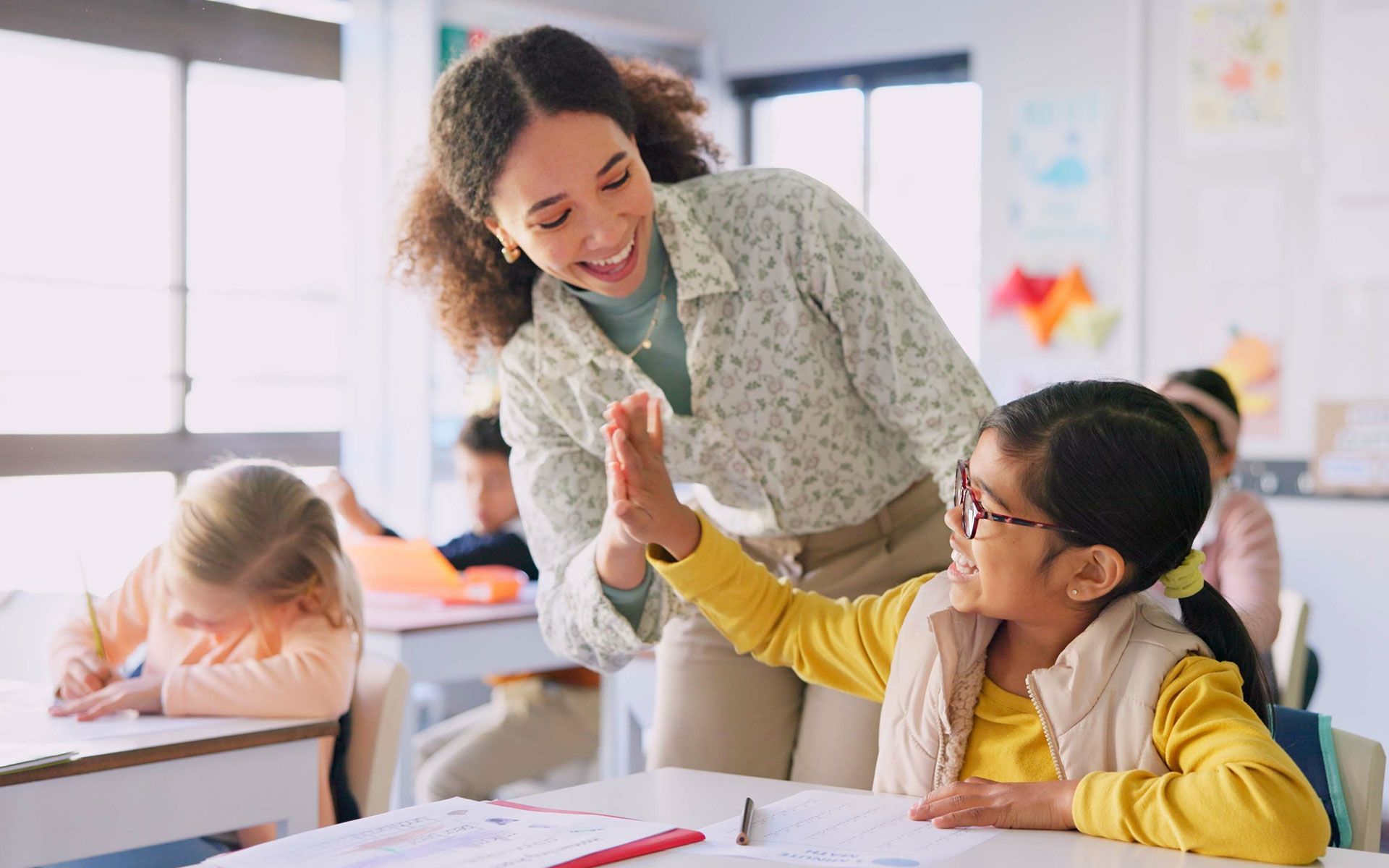 Teacher high-fives a student in a classroom. The student is wearing glasses and smiles. Others are seated at desks.