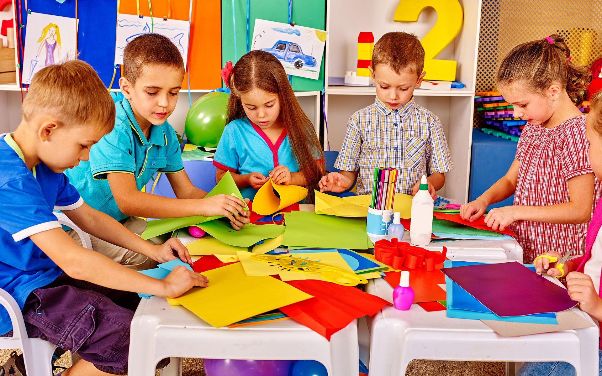Children at a table doing a craft project, using colorful paper and glue in a brightly lit room.