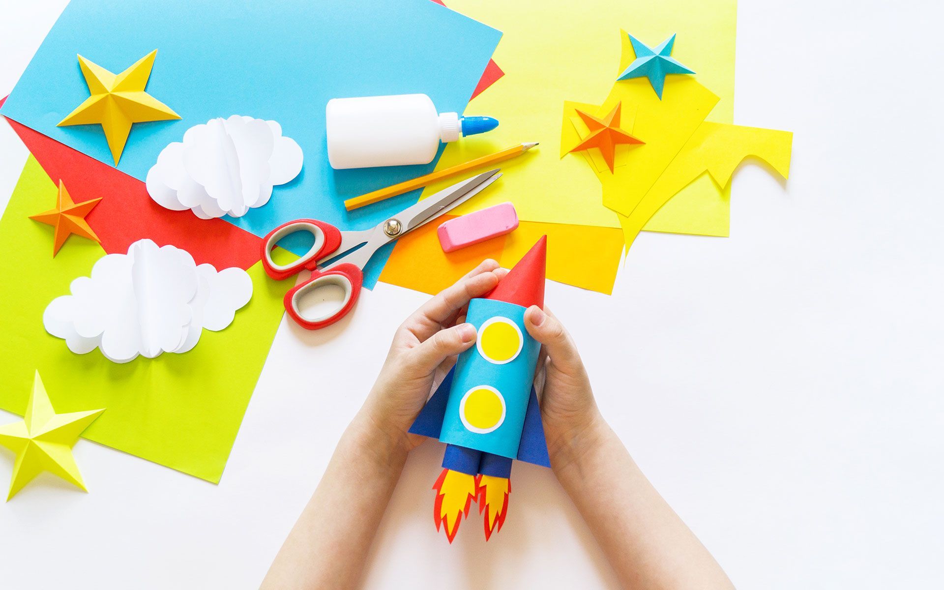 Hands hold a paper rocket ship with craft supplies: stars, clouds, and colorful paper.