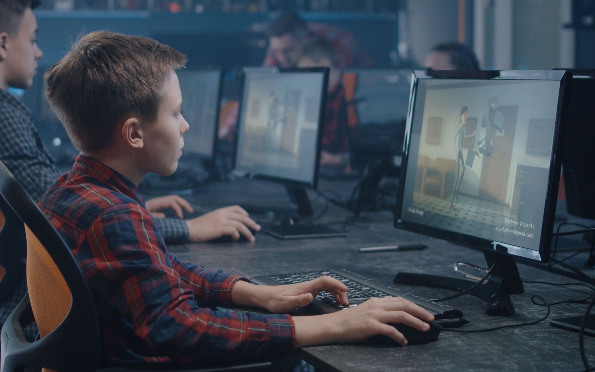 Boy in plaid shirt playing a video game at a computer desk, other people in the background.