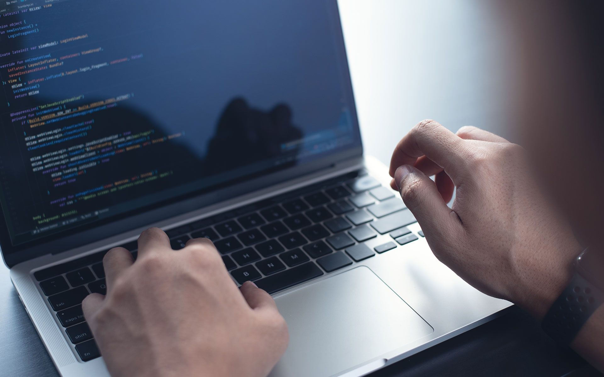 Person typing code on a laptop, close-up of hands and screen. Dark blue code visible with a gray laptop.