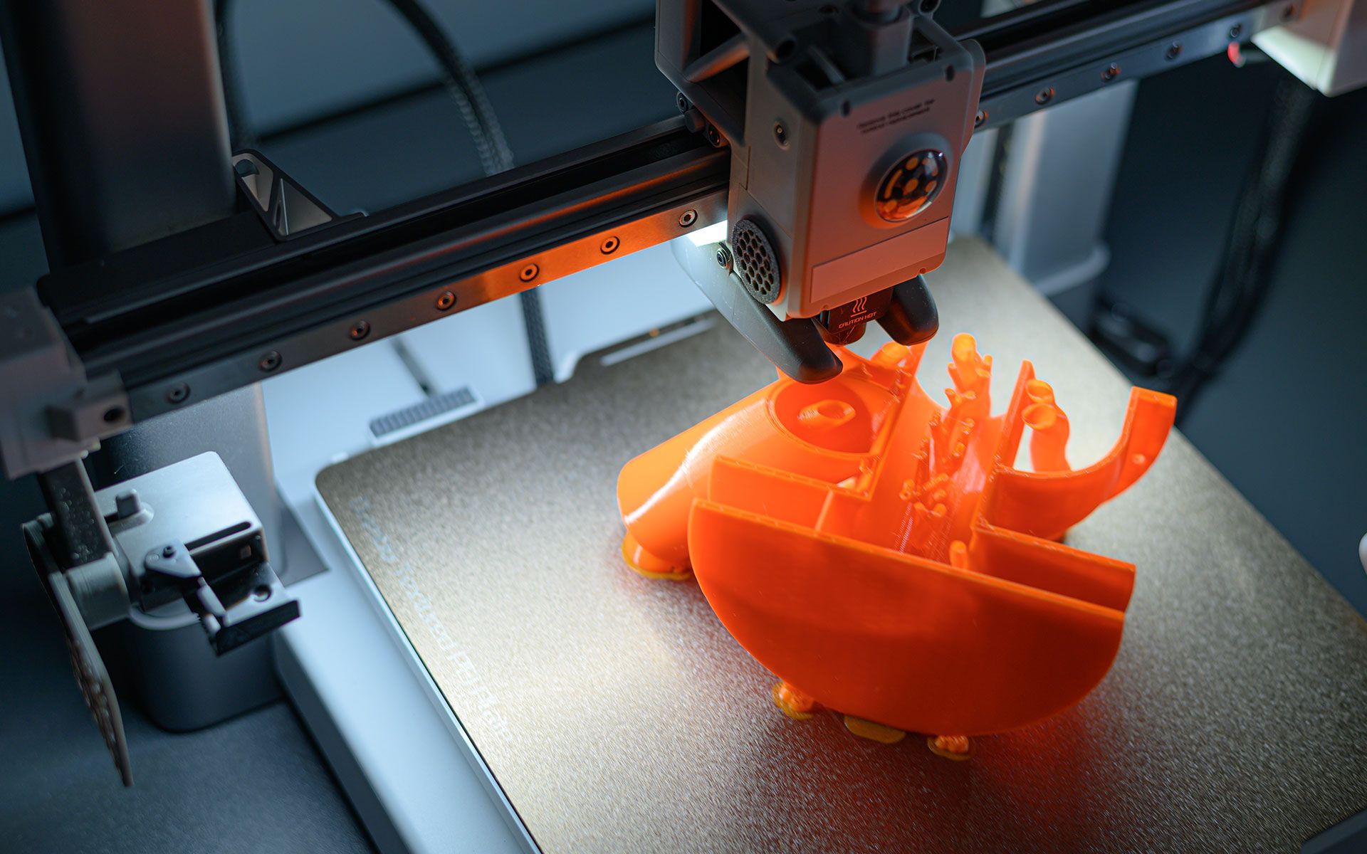 3D printer depositing orange plastic layers to create an object on a silver platform.