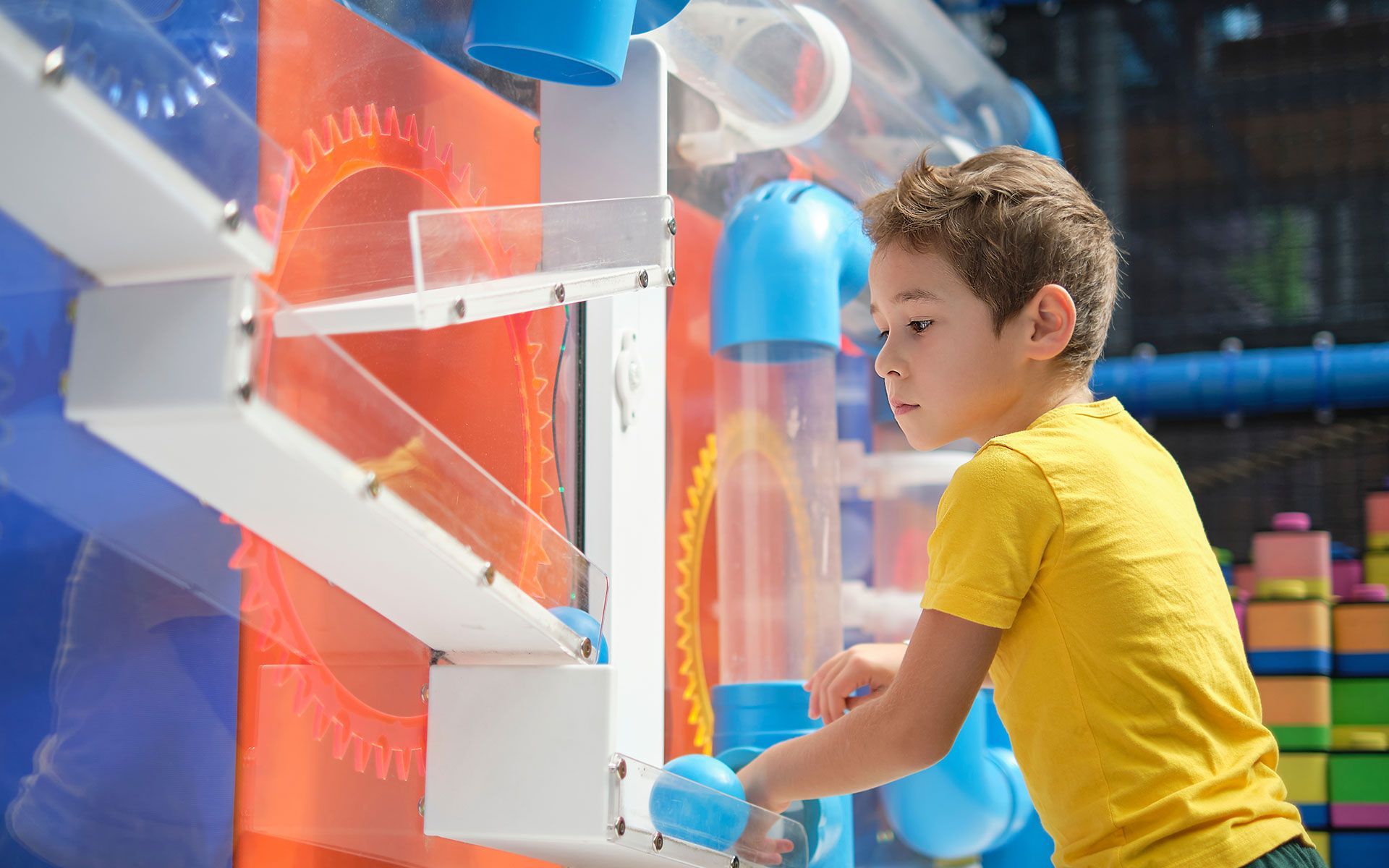 Boy in yellow shirt plays with a colorful ball-run toy at an indoor play area.