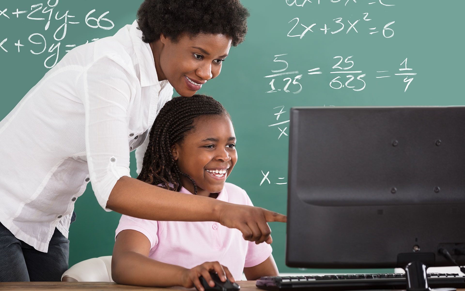 Teacher points at computer screen with student, both smiling in front of a chalkboard.