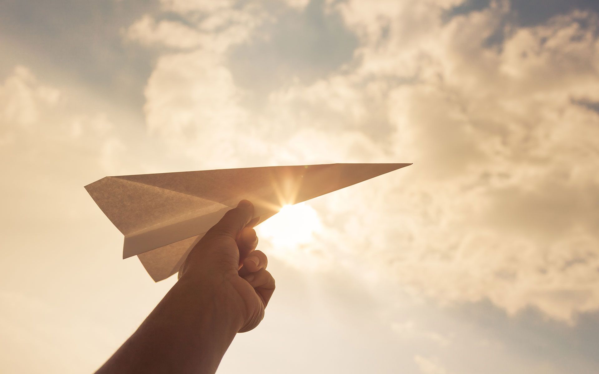 Hand releasing a paper airplane toward the sun in a cloudy sky.