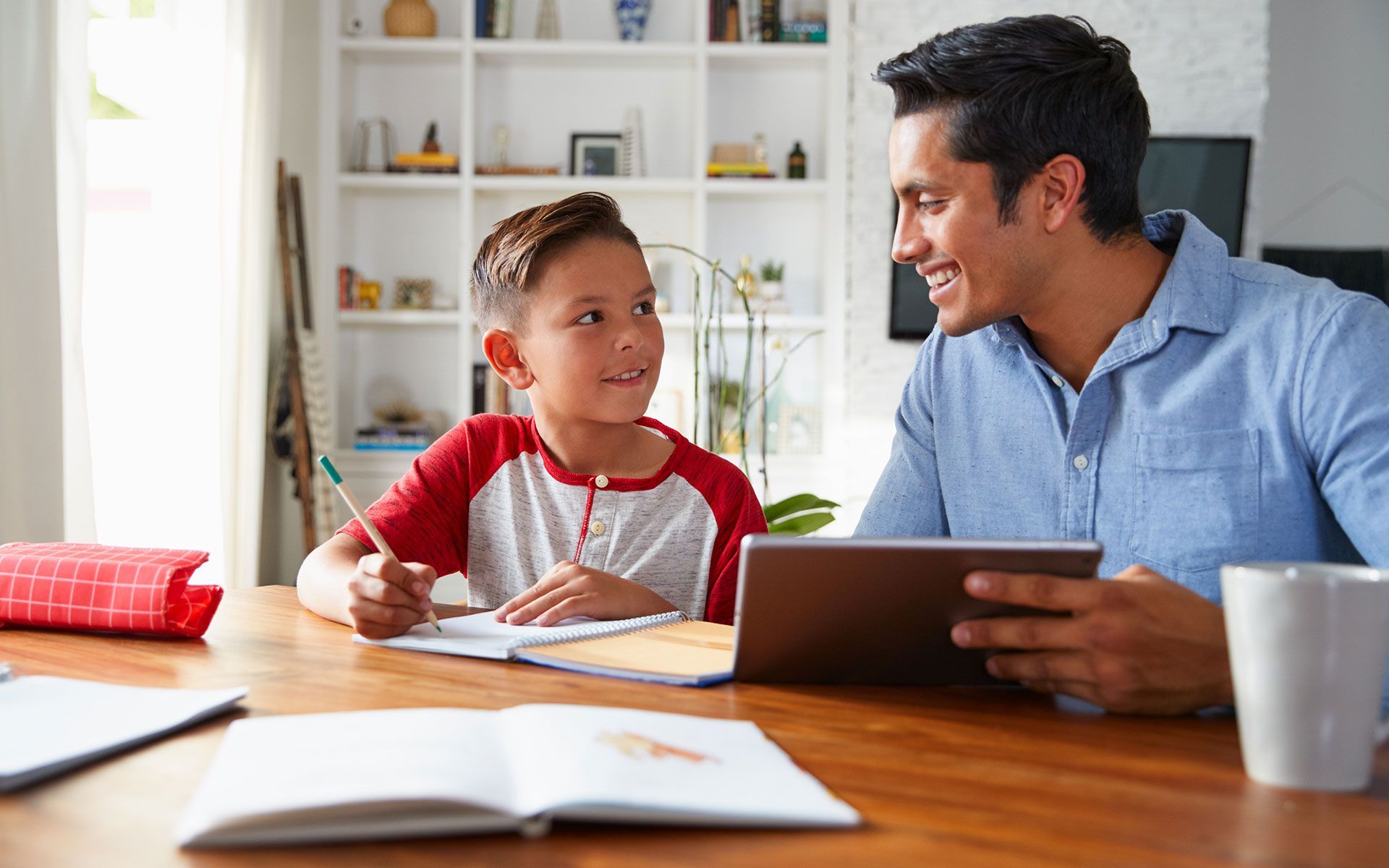 Man with a tablet assists a child doing homework at a wooden table in a well-lit room.