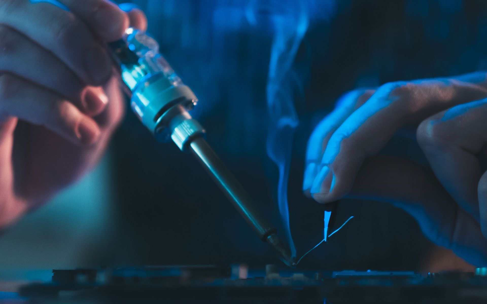 Hands soldering electronics with a hot iron, blue light.