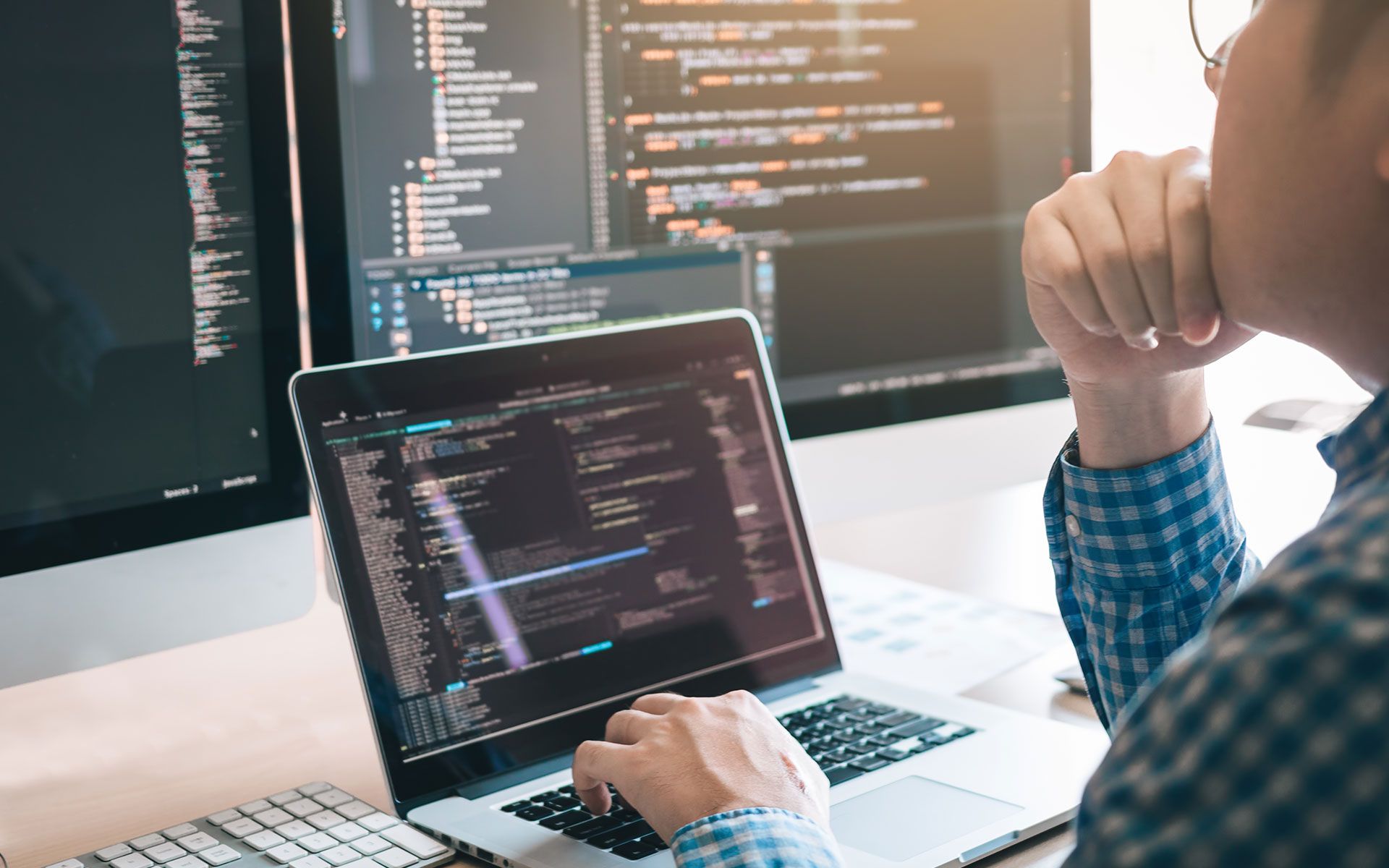 Person coding on a laptop with multiple computer screens displaying code. The person is deep in thought.
