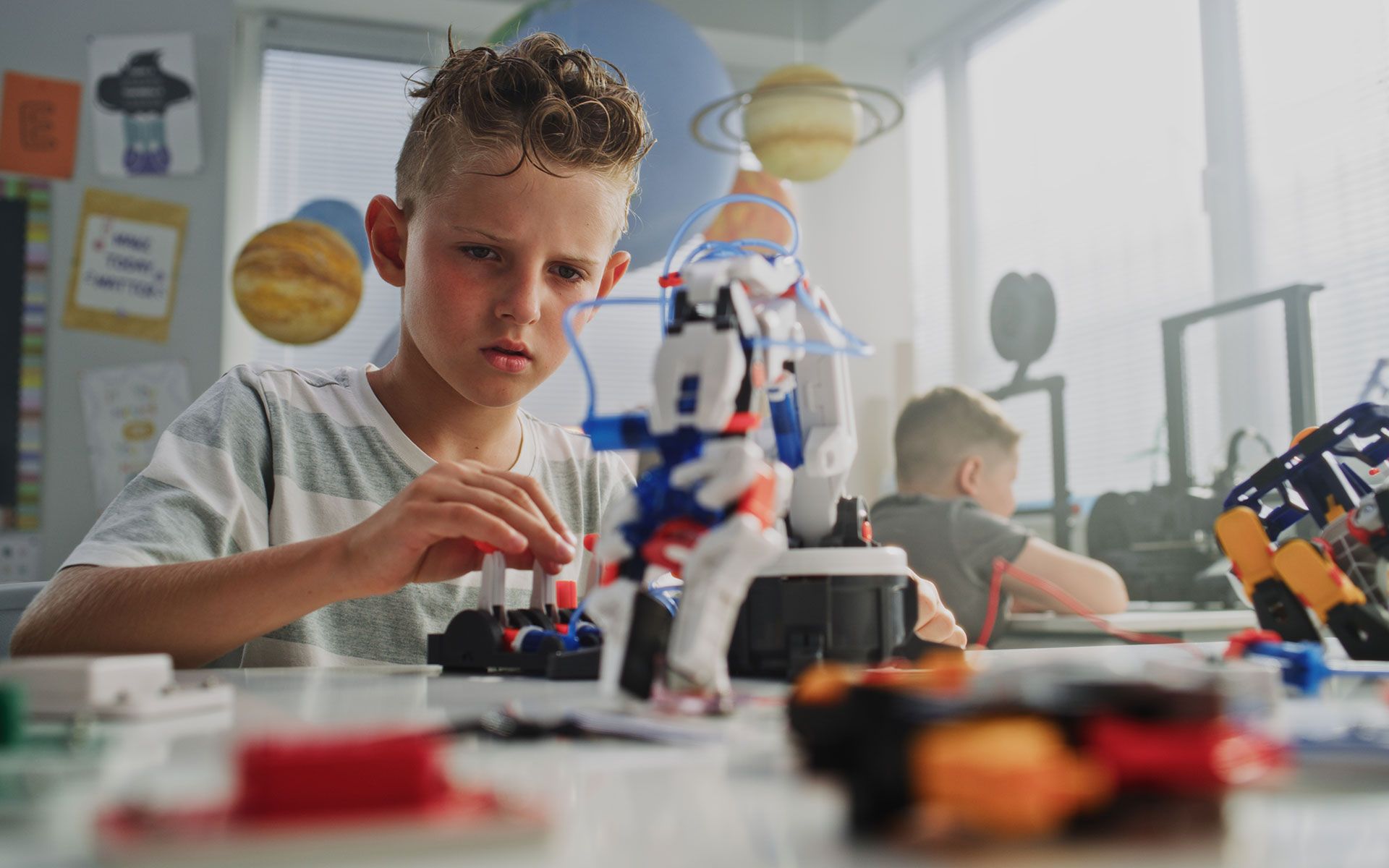 Boy focused on building a robot at a desk, classroom setting, other children in background.