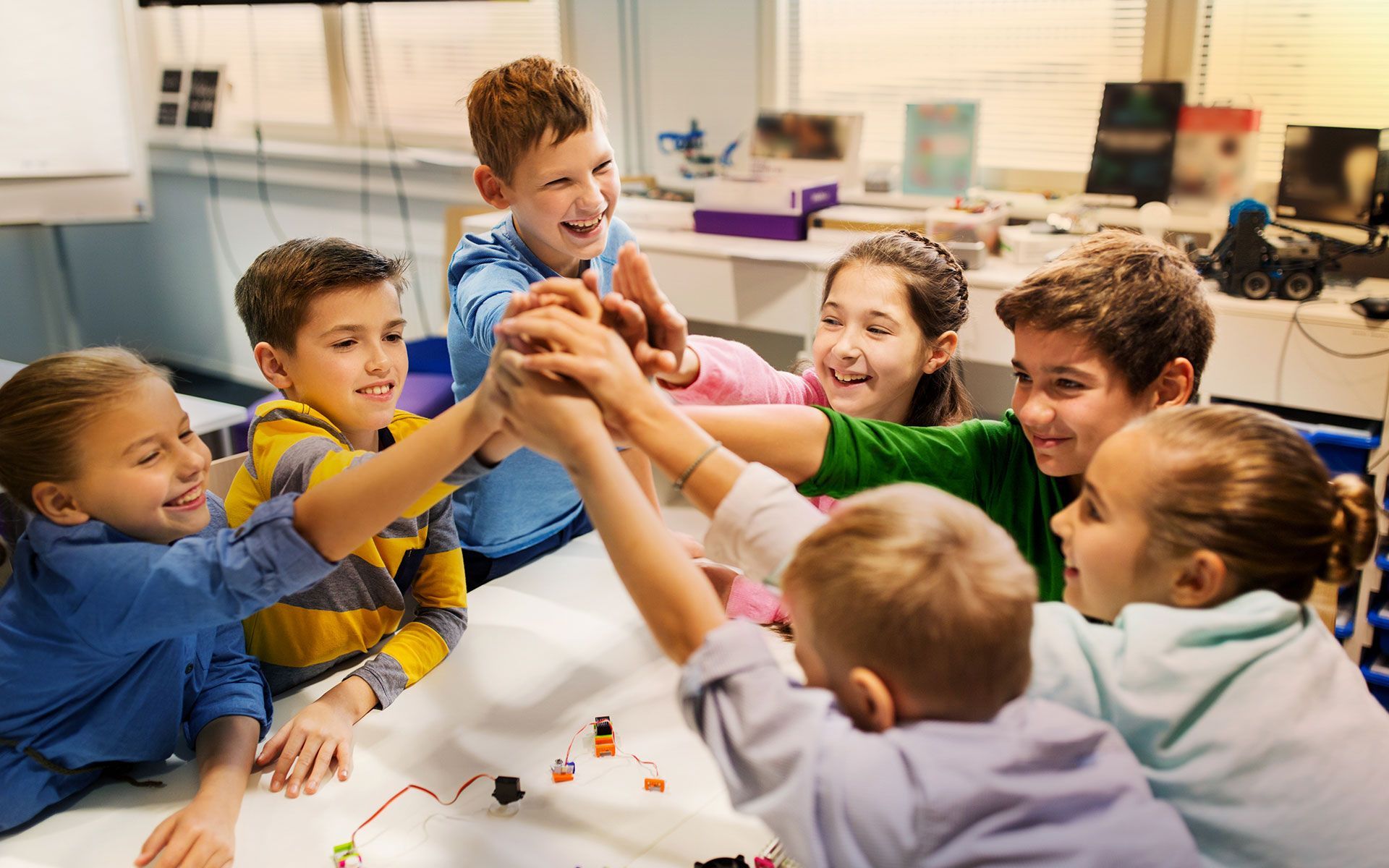 Children in classroom huddle, hands together, smiling.