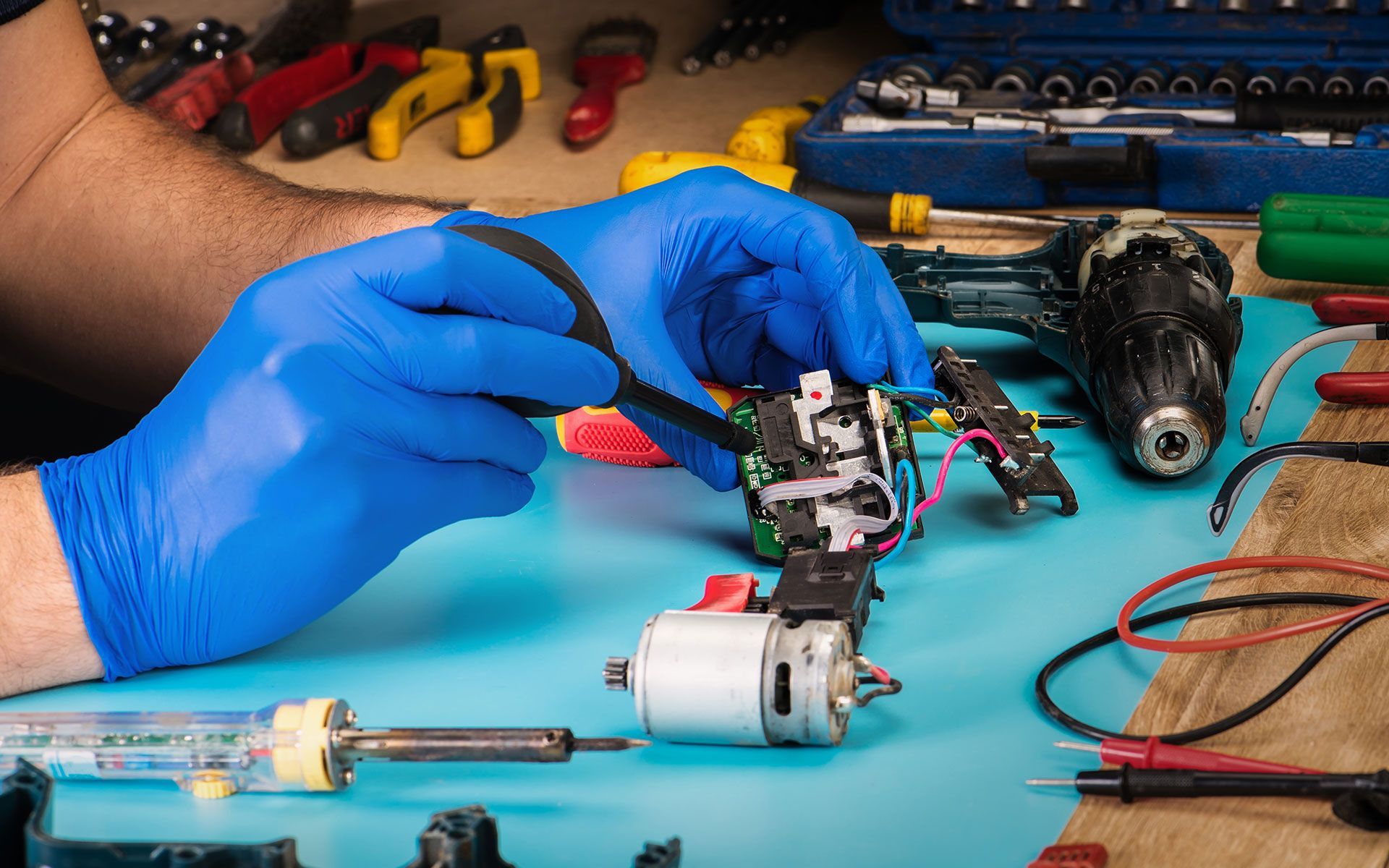 Hands in blue gloves using a screwdriver to repair a small motor on a blue surface.