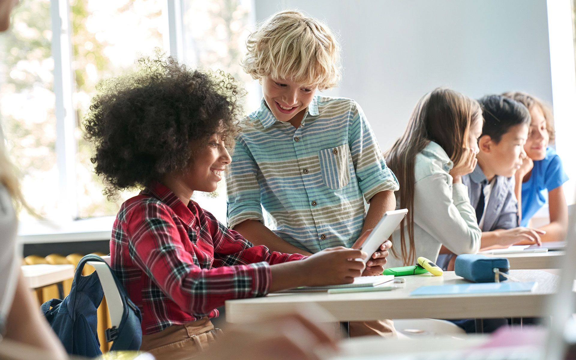 Two students at a desk looking at a tablet, smiling. Other students in classroom.