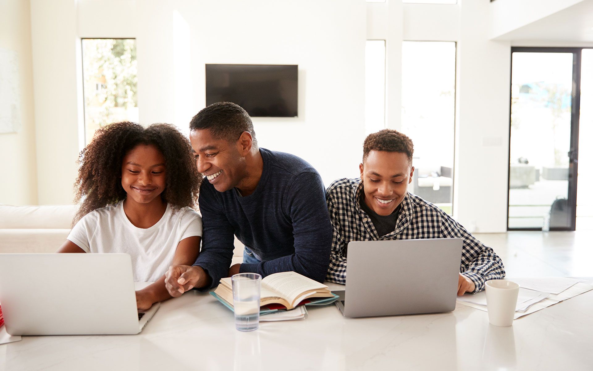 Father assisting two children with their laptops at a counter, smiling, in a bright home.