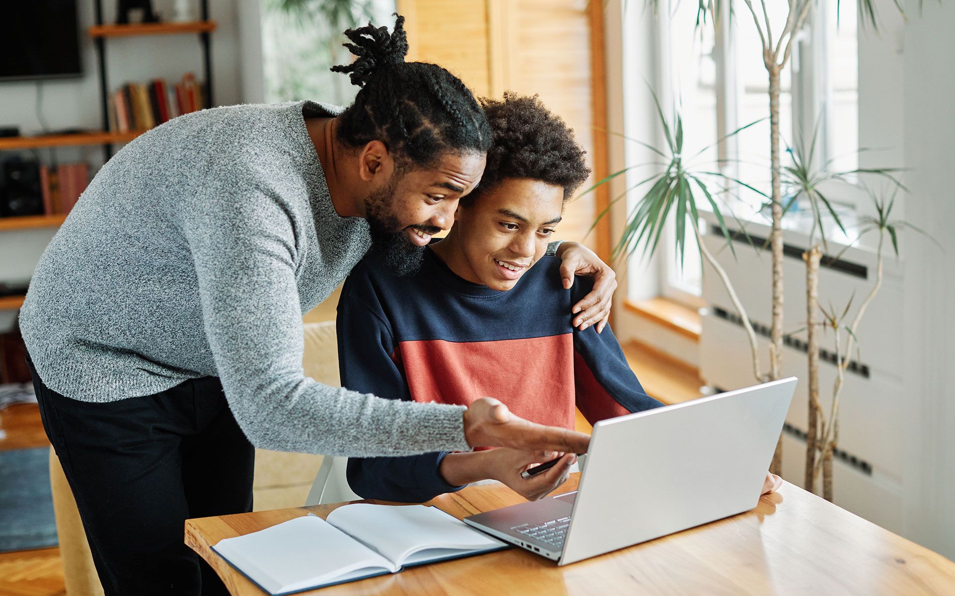 Man helping teenager with laptop, pointing at screen. Living room setting with plant.