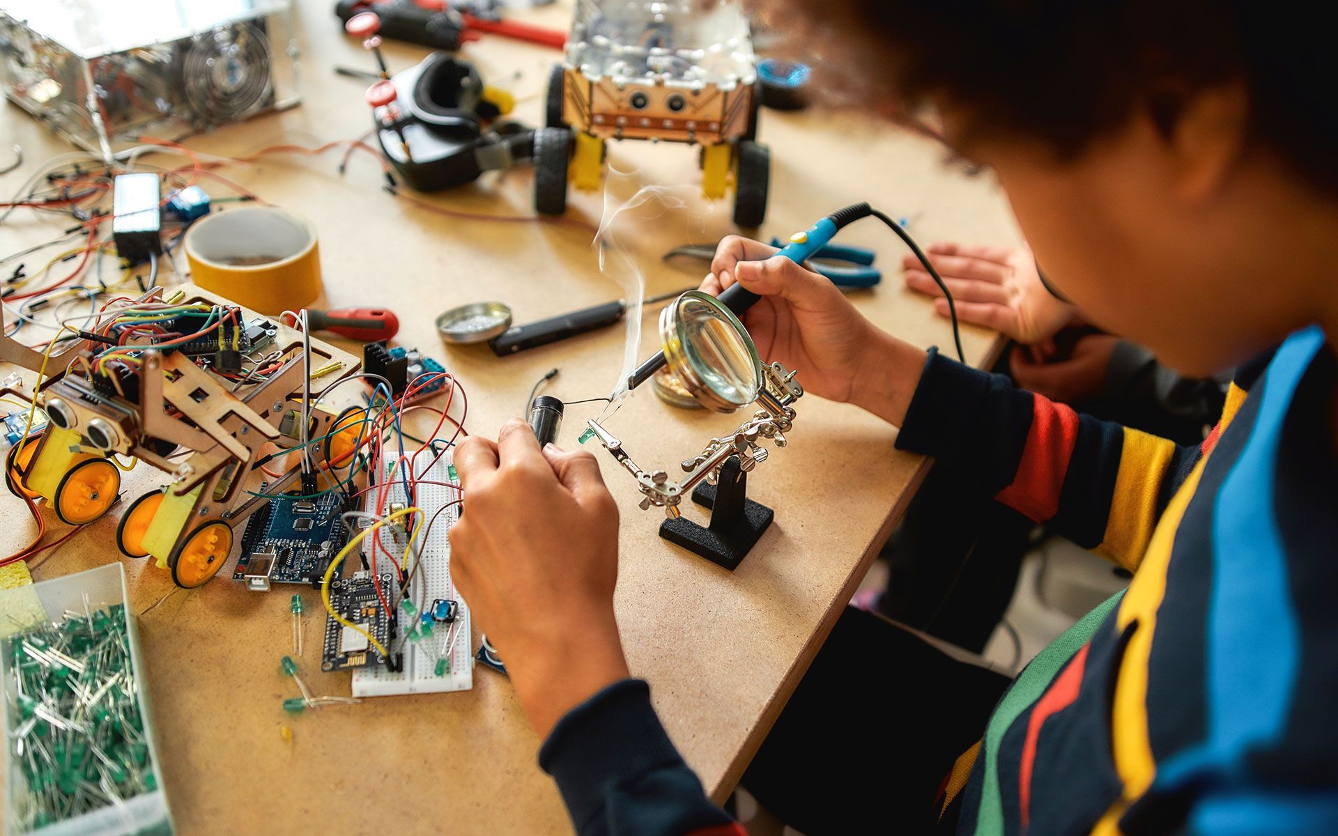 Person soldering wires with magnifying glass at a workbench with robot parts and tools.