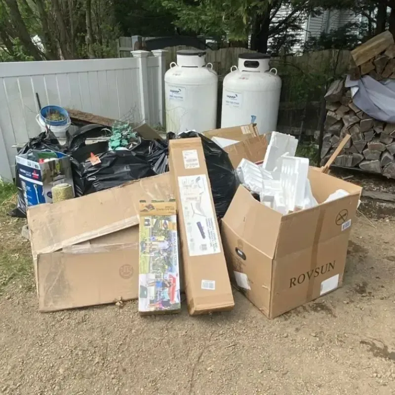 Pile of trash and cardboard boxes outdoors, near propane tanks and firewood.