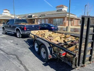 Truck towing a trailer loaded with firewood in a parking lot, in front of a brick building.