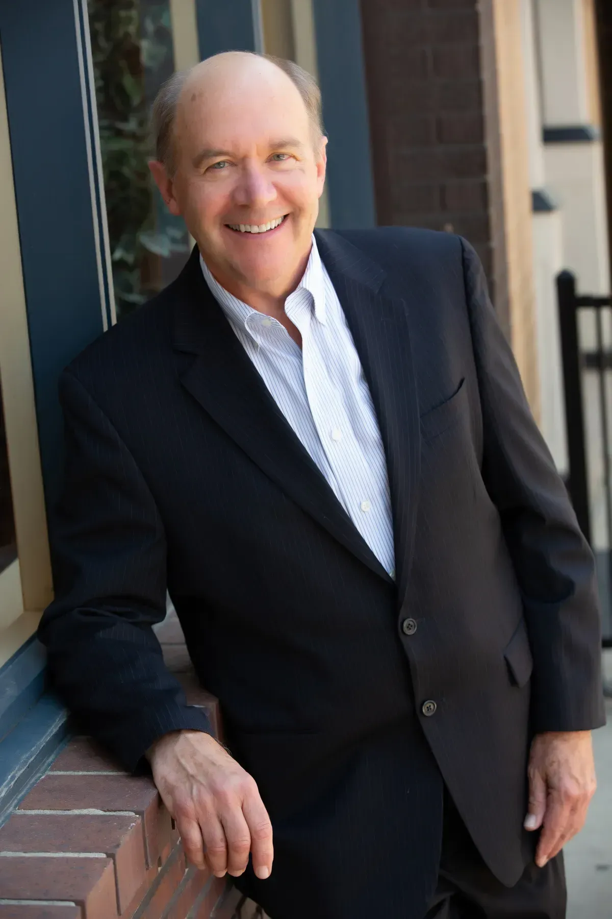 A man in a suit is leaning against a brick wall and smiling