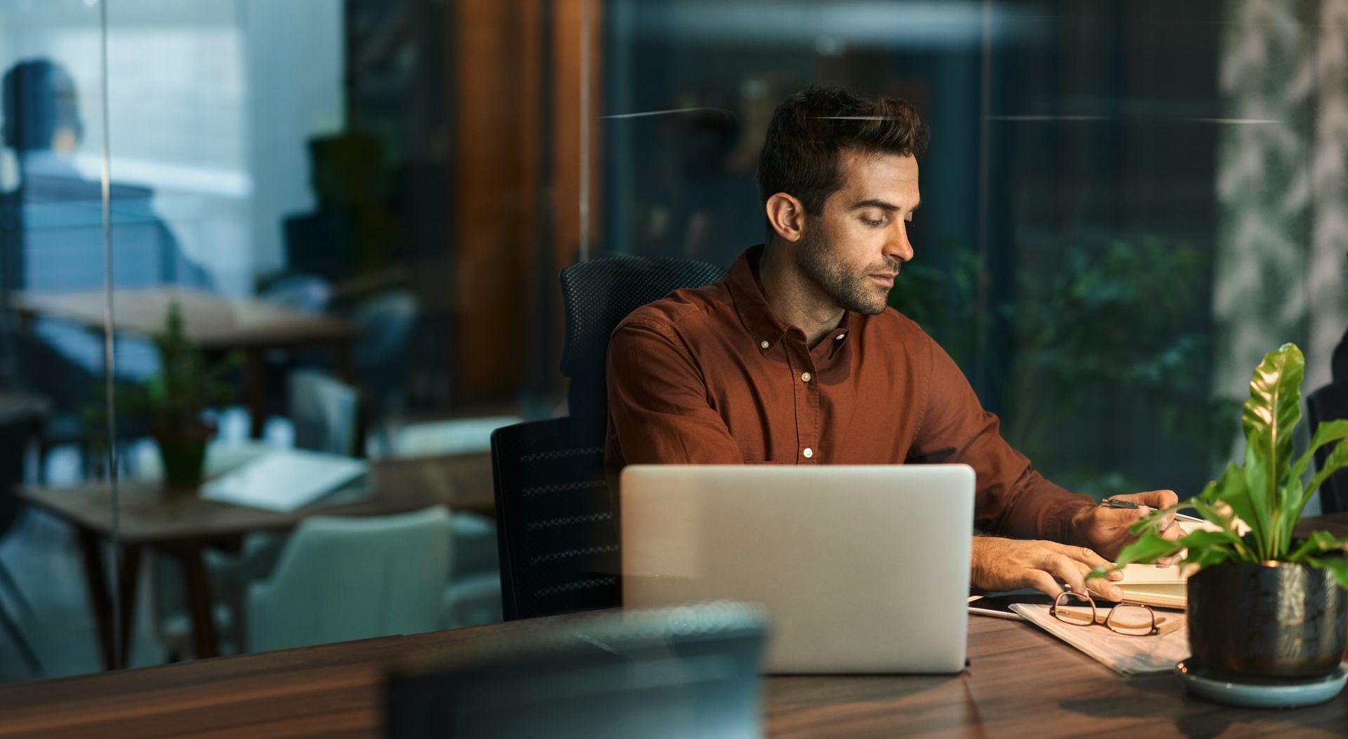 Man working on laptop at desk in office, focused. Dark room, warm lighting.