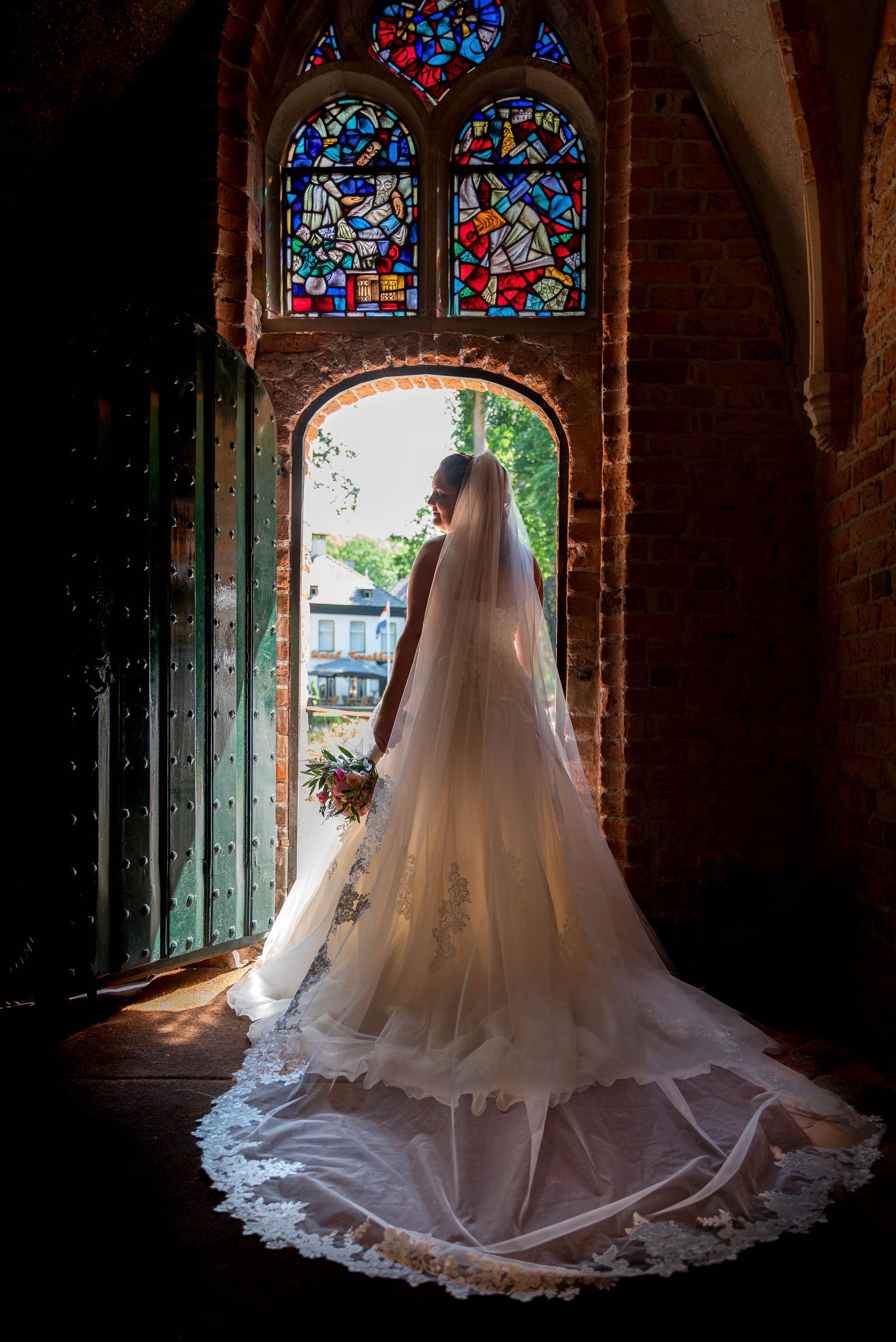 Een bruid in een trouwjurk en sluier staat voor een glas-in-loodraam.
