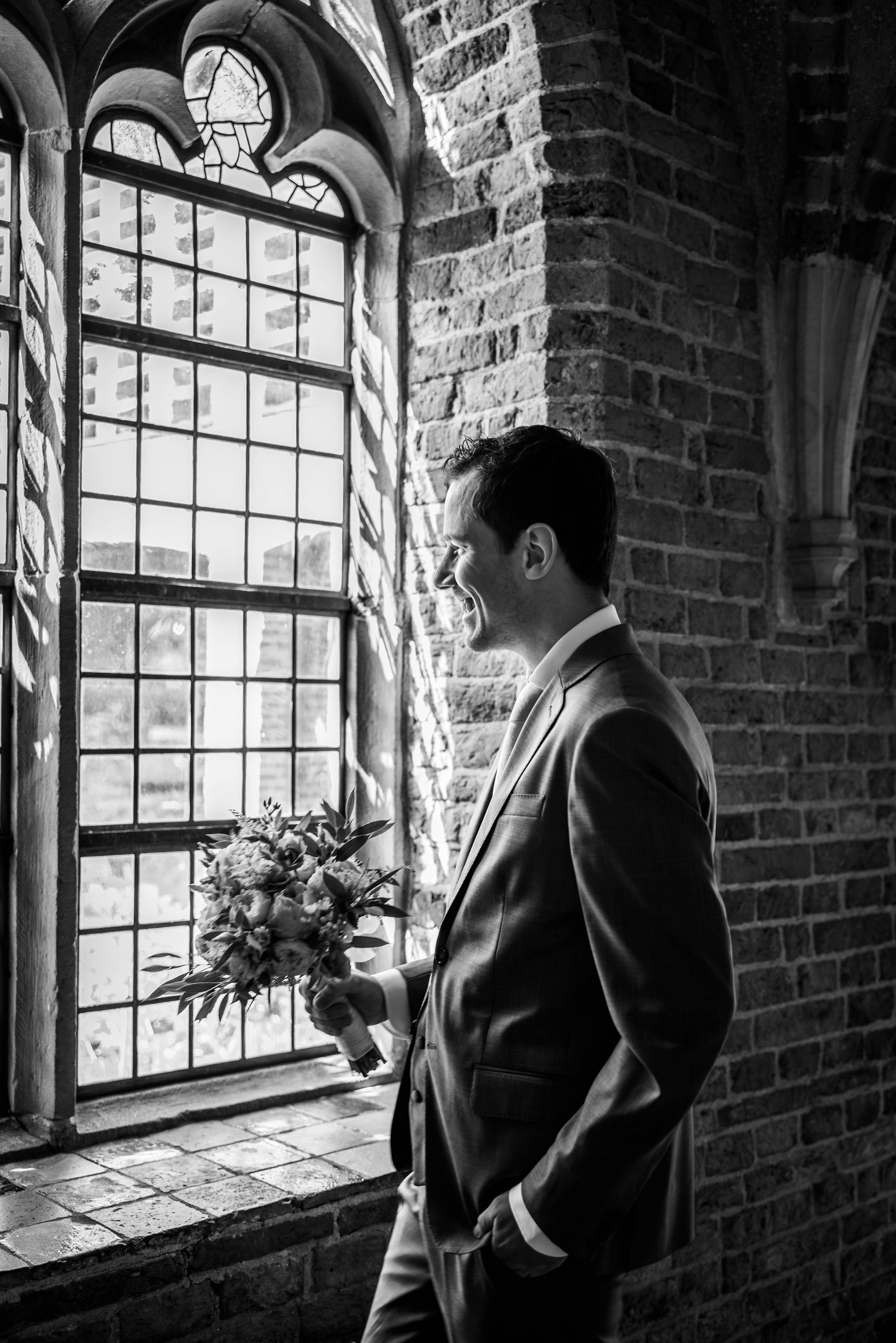 Een man in pak staat voor een raam met een boeket bloemen in zijn handen.