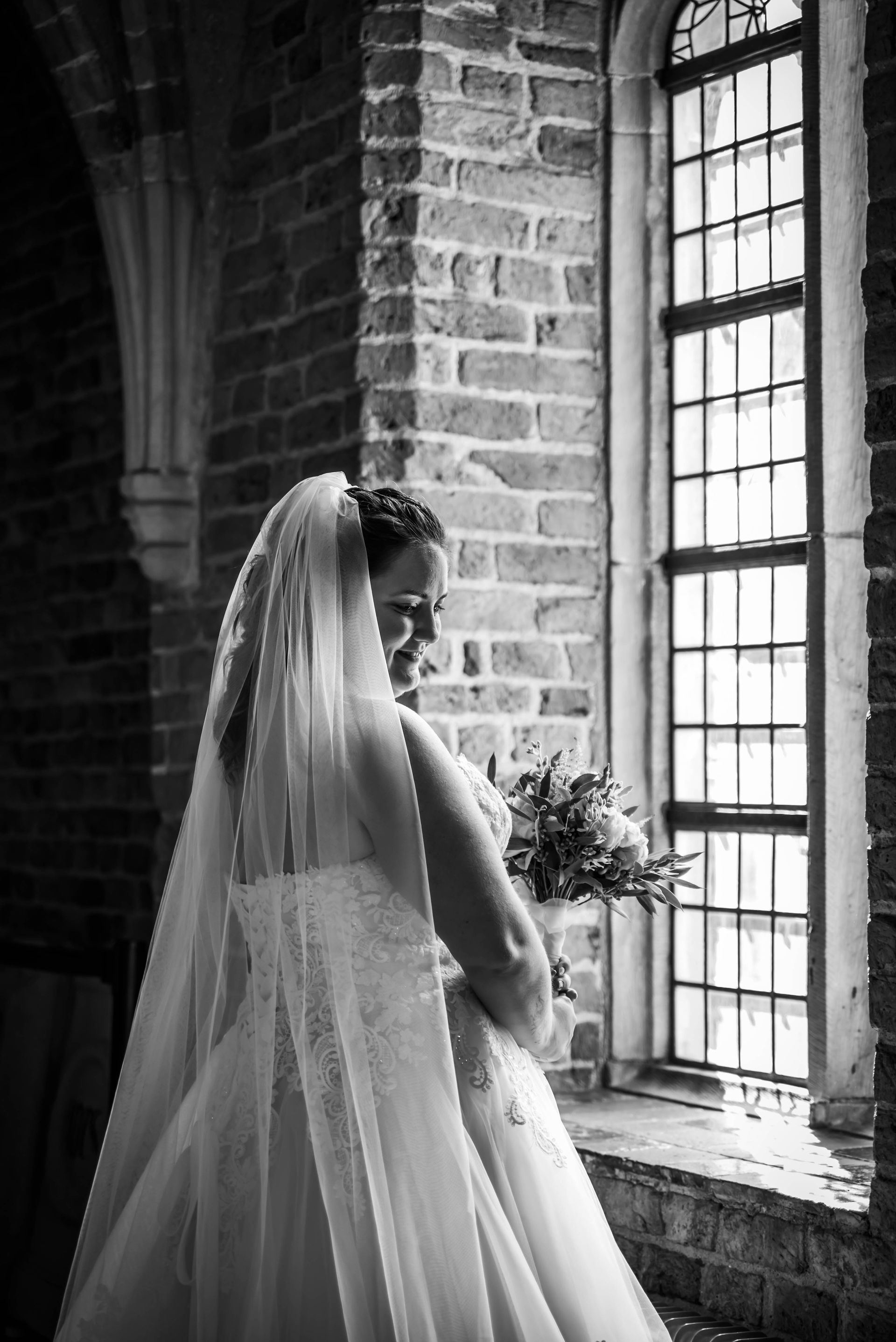 Een zwart-witfoto van een bruid in een trouwjurk die voor een raam staat.