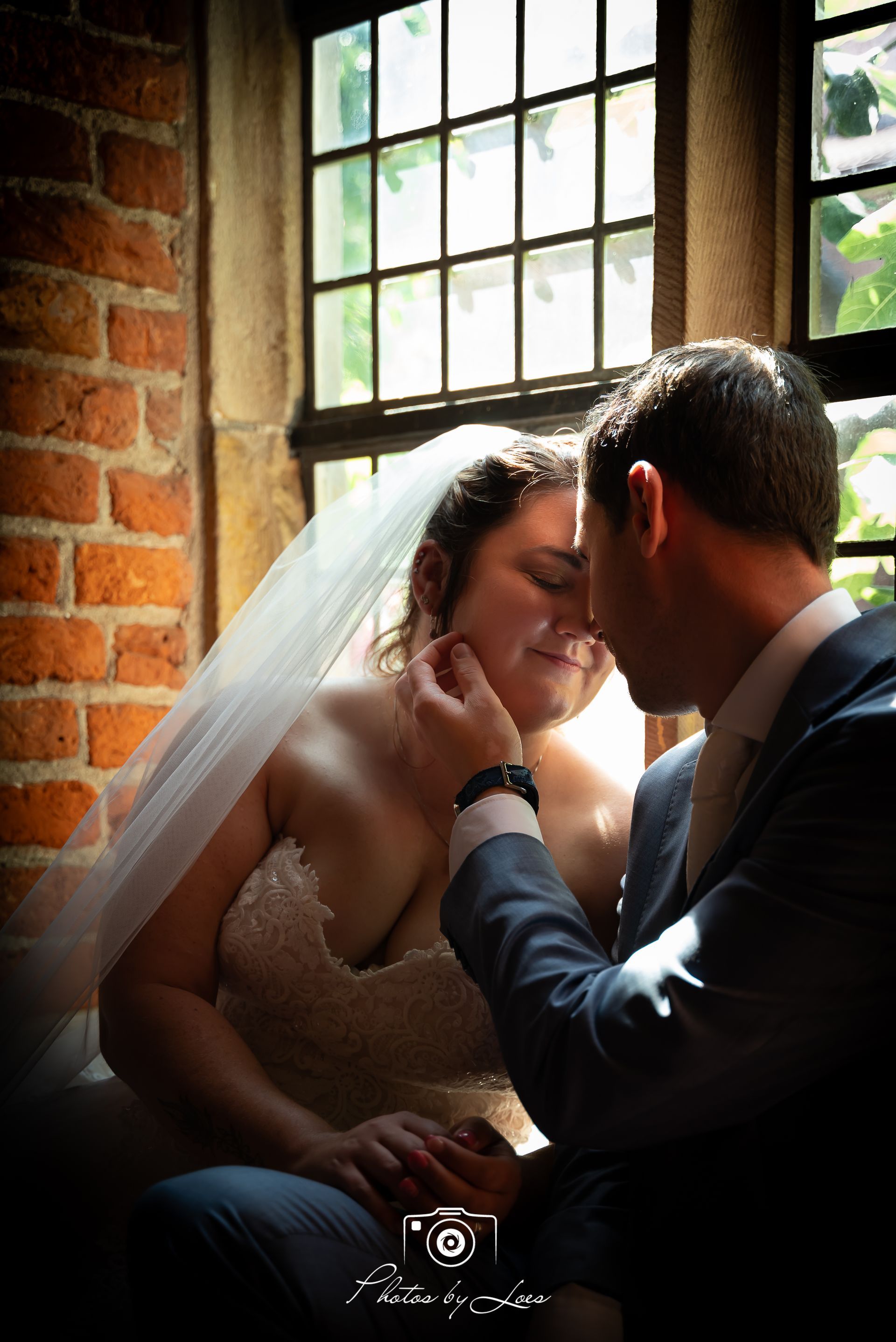 Een bruid en bruidegom kussen elkaar voor een raam.