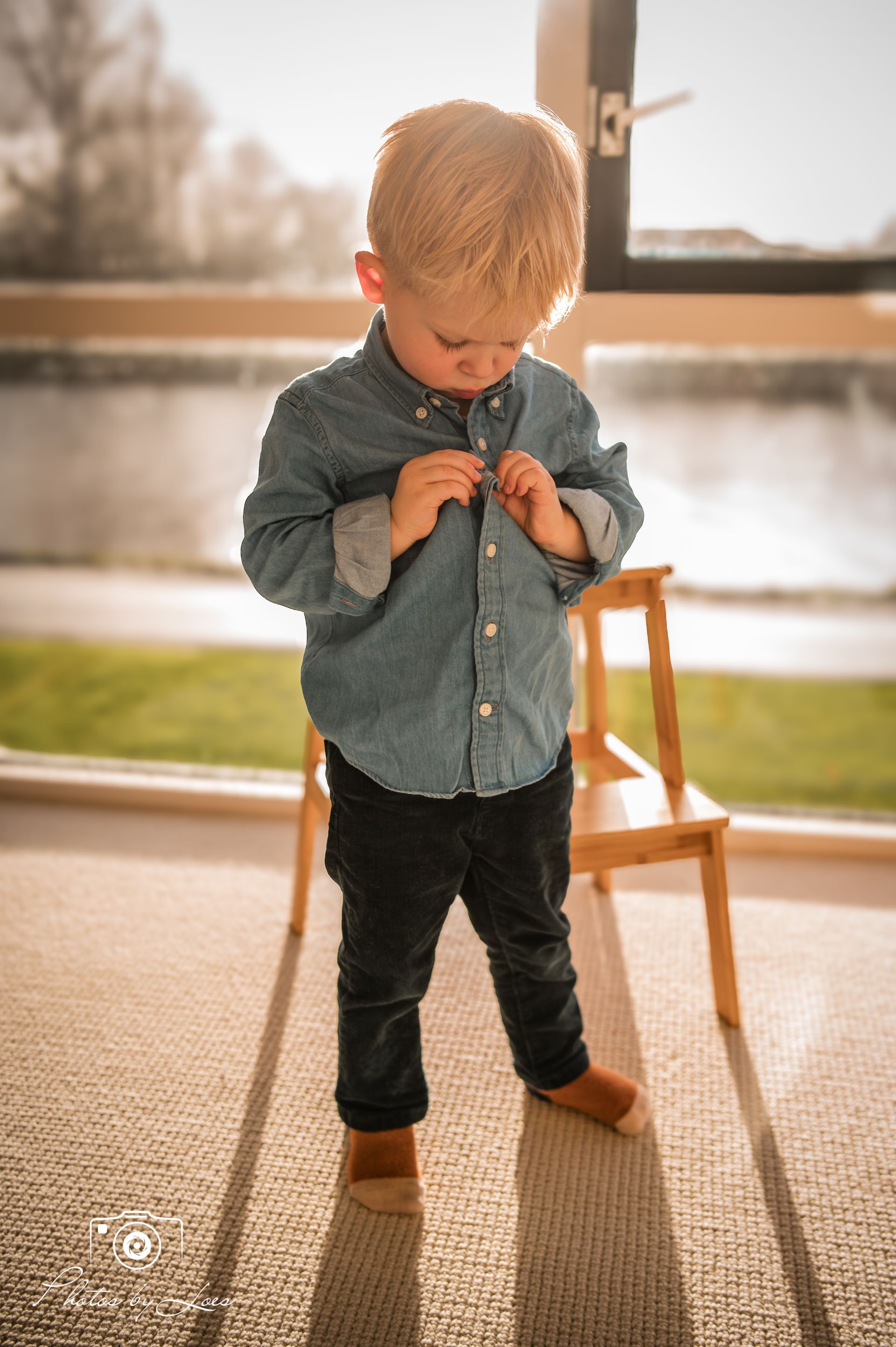 Een jonge jongen staat voor een raam en trekt zijn shirt recht.