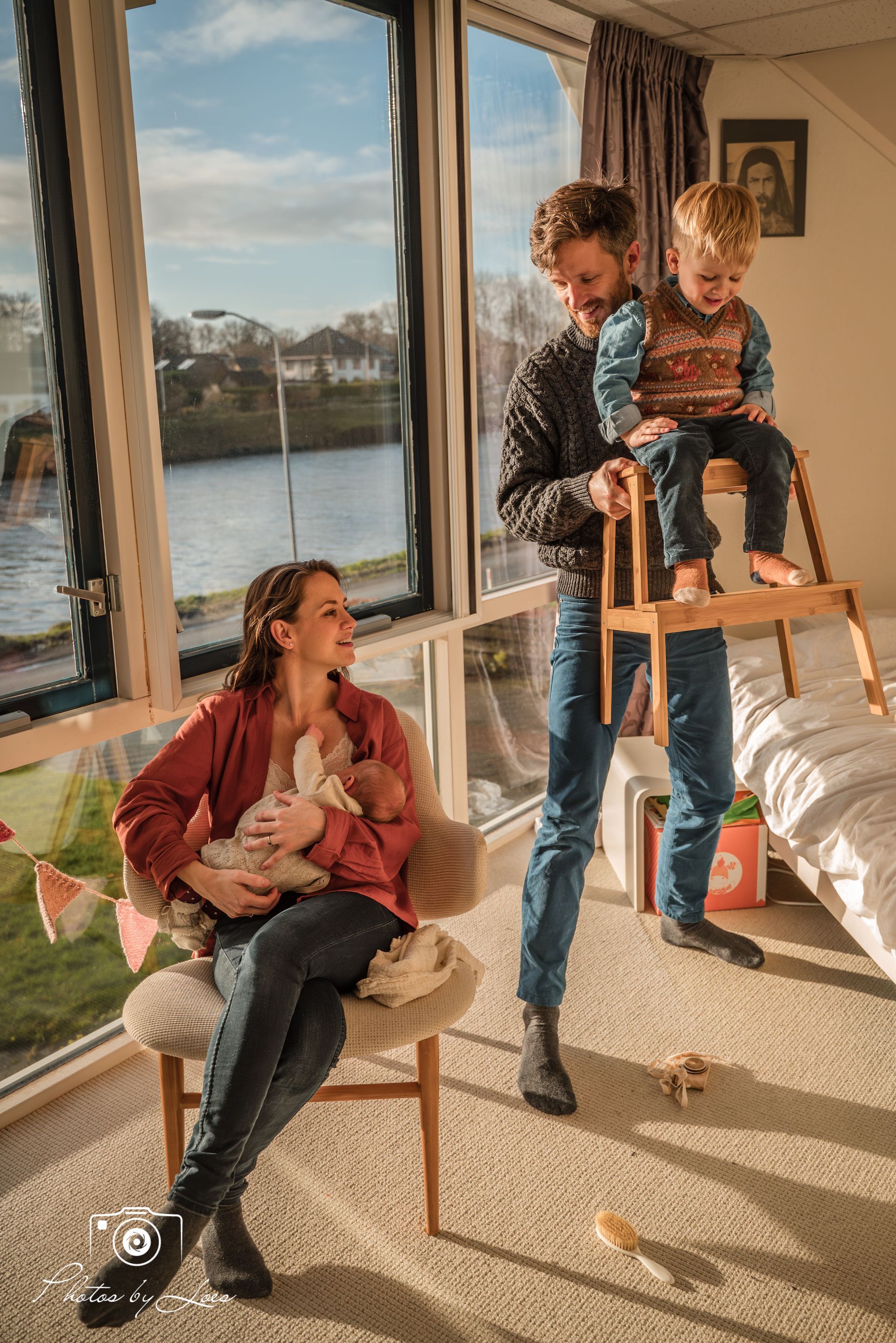 Een vrouw zit op een stoel met een baby in haar armen, terwijl een man een kind op een ladder vasthoudt.