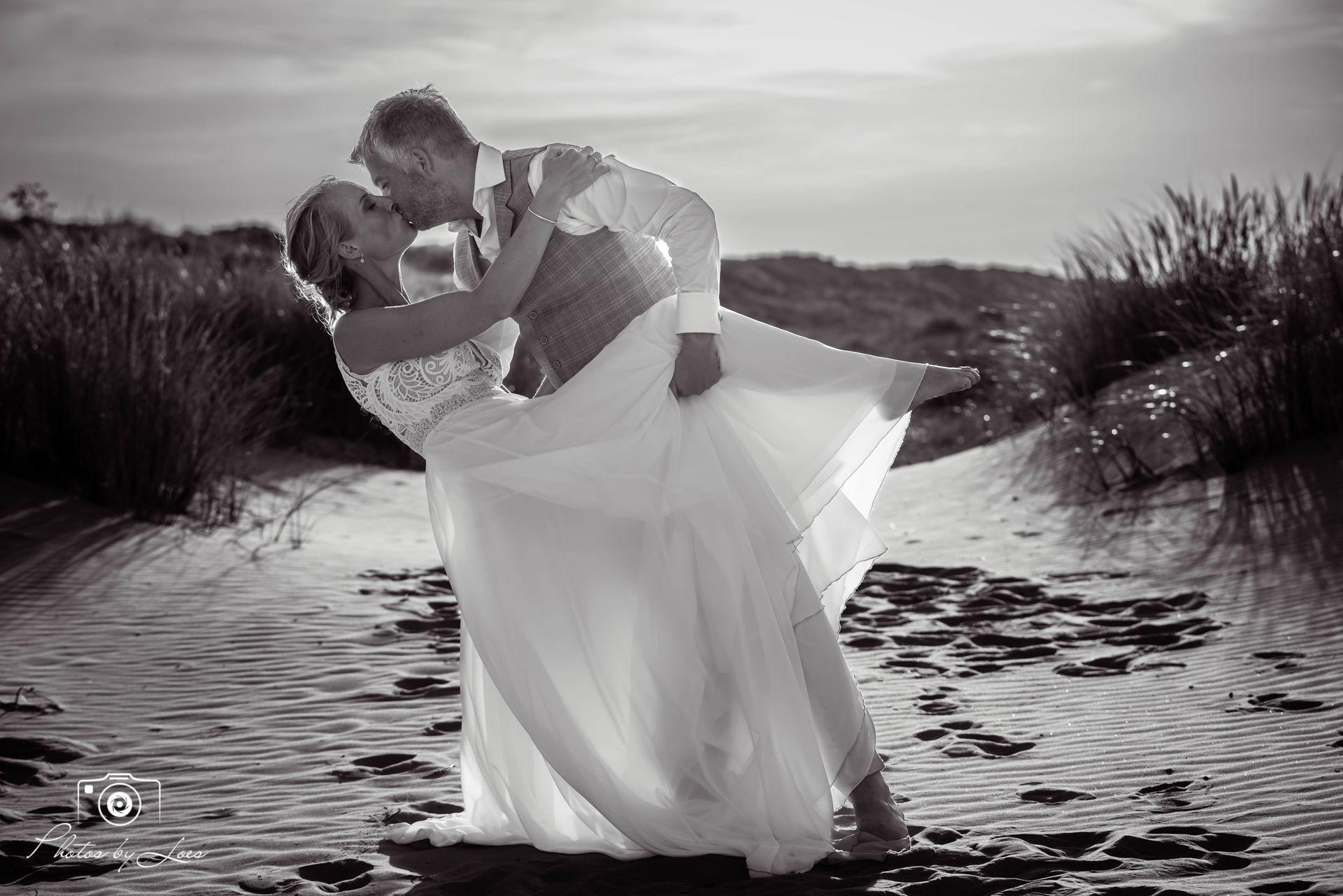 Op een zwart-witfoto is een bruid en bruidegom aan het kussen op het strand.