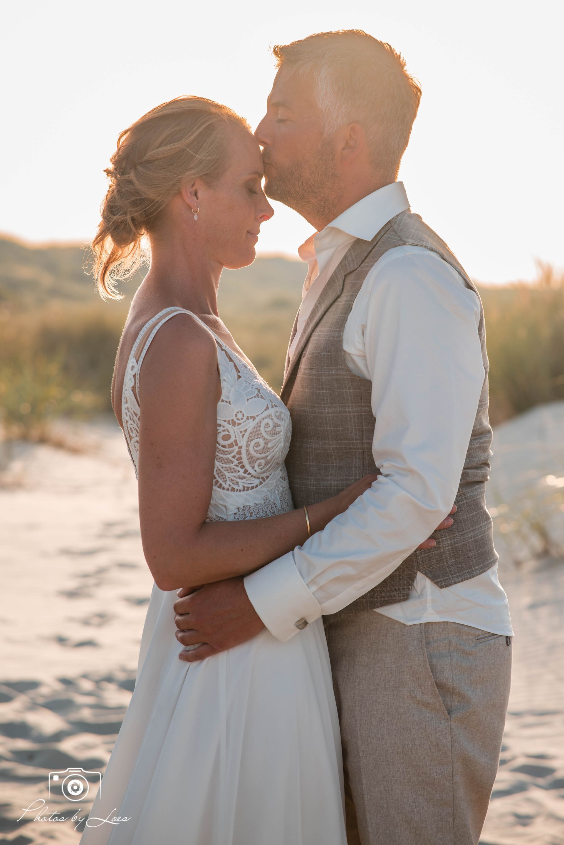 Een bruid en bruidegom kussen elkaar op het strand bij zonsondergang.