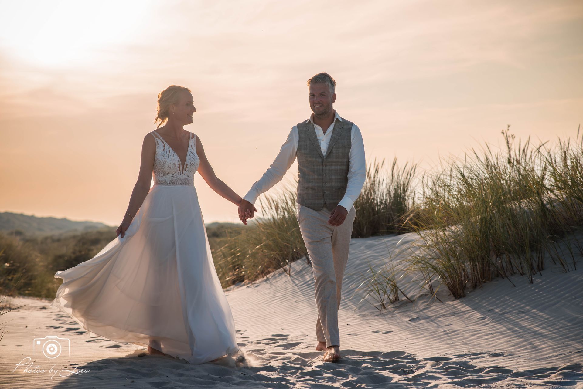 Een bruid en bruidegom lopen hand in hand over een zandpad.