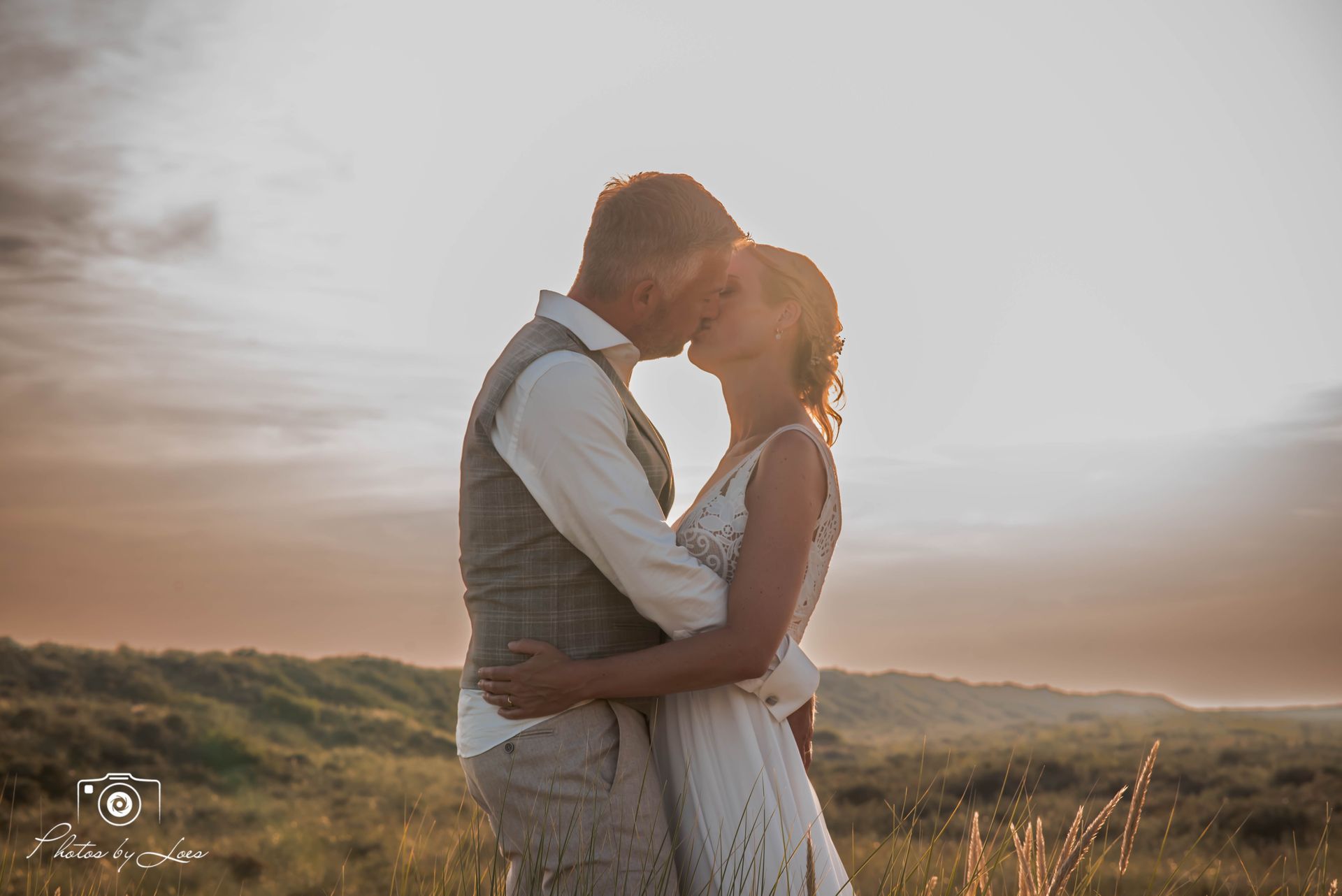 Een bruid en bruidegom kussen elkaar in een veld bij zonsondergang.