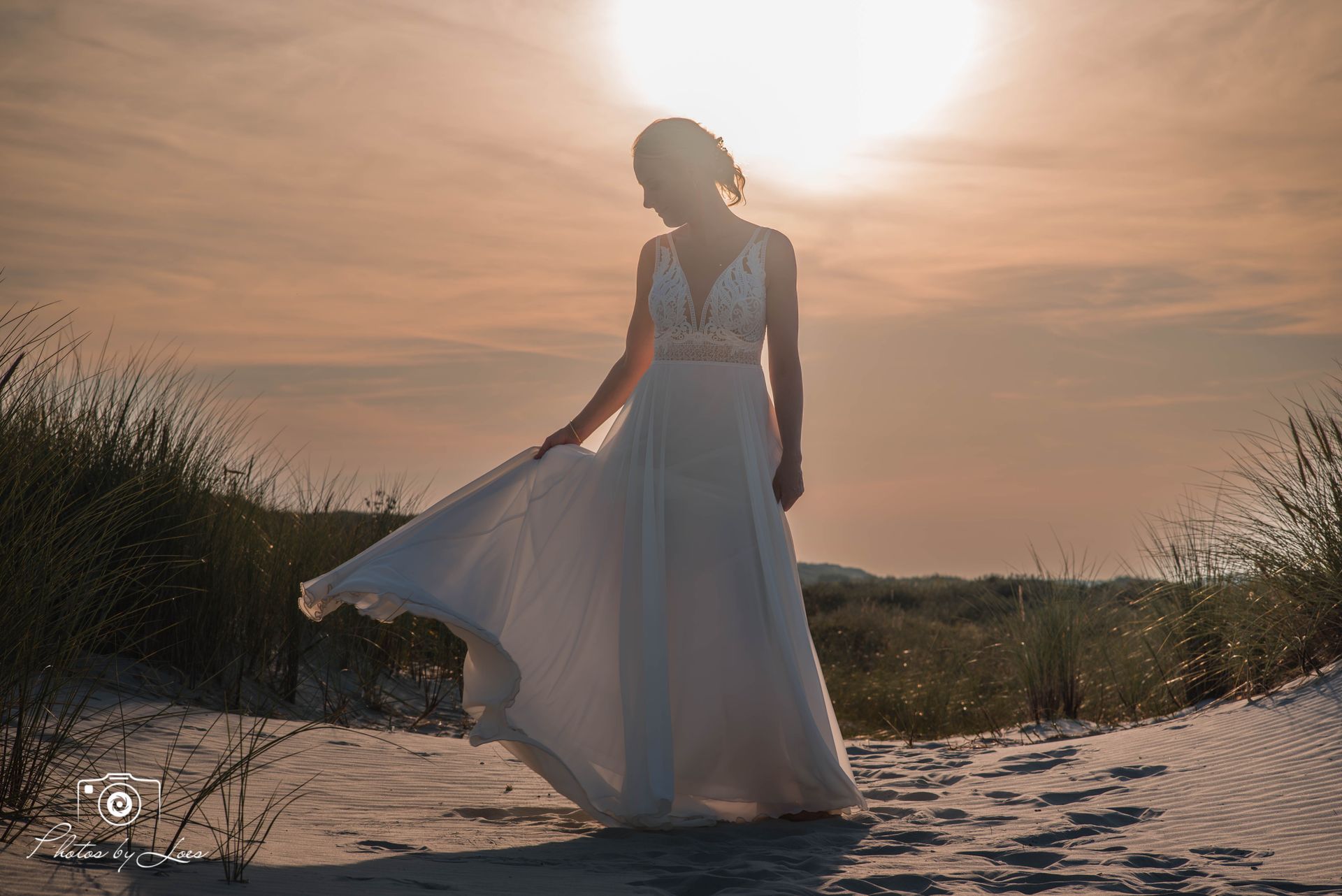Een vrouw in een trouwjurk staat op een zandduin bij zonsondergang.