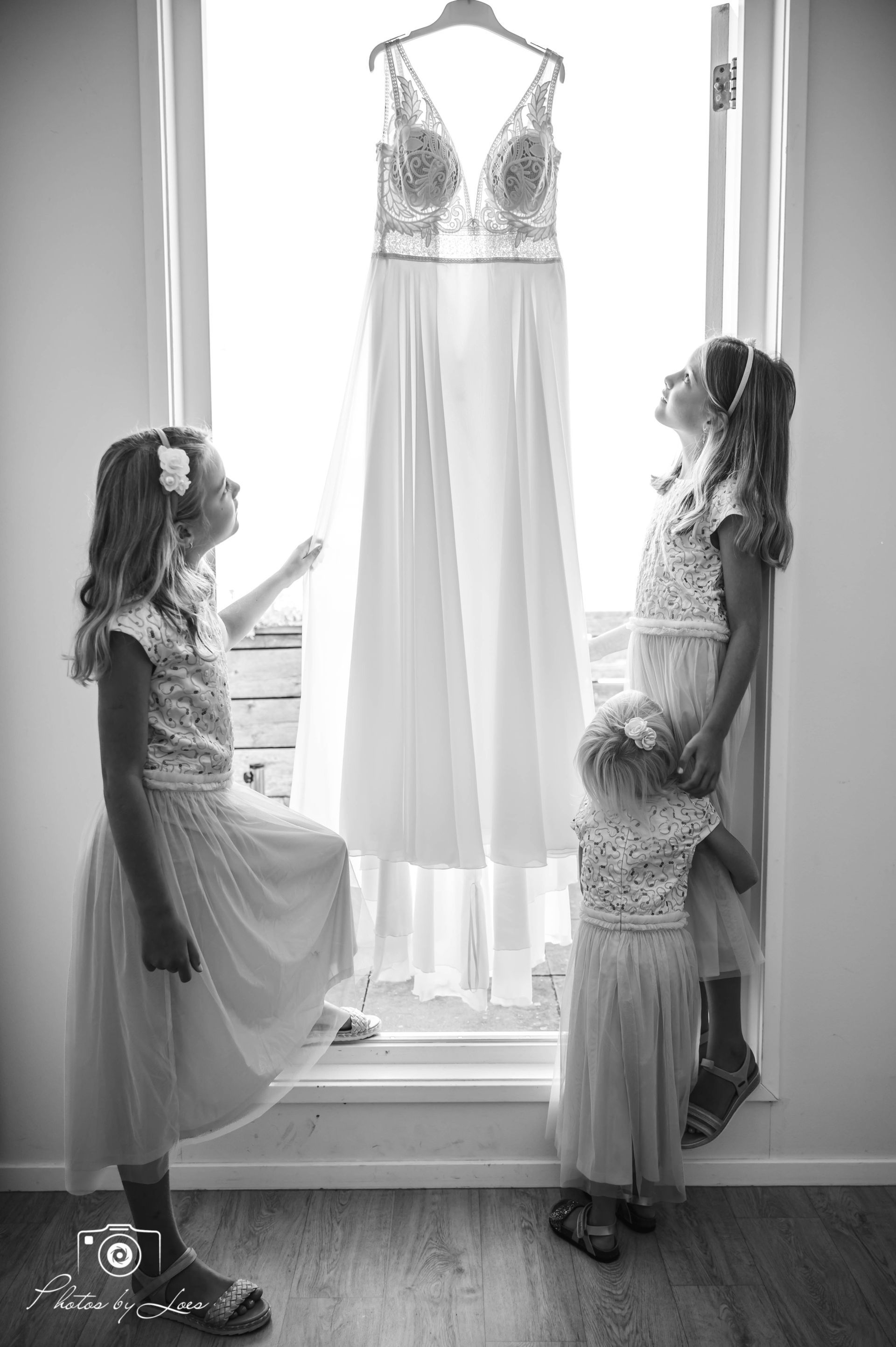 Drie kleine meisjes kijken naar een trouwjurk die in een raam hangt.