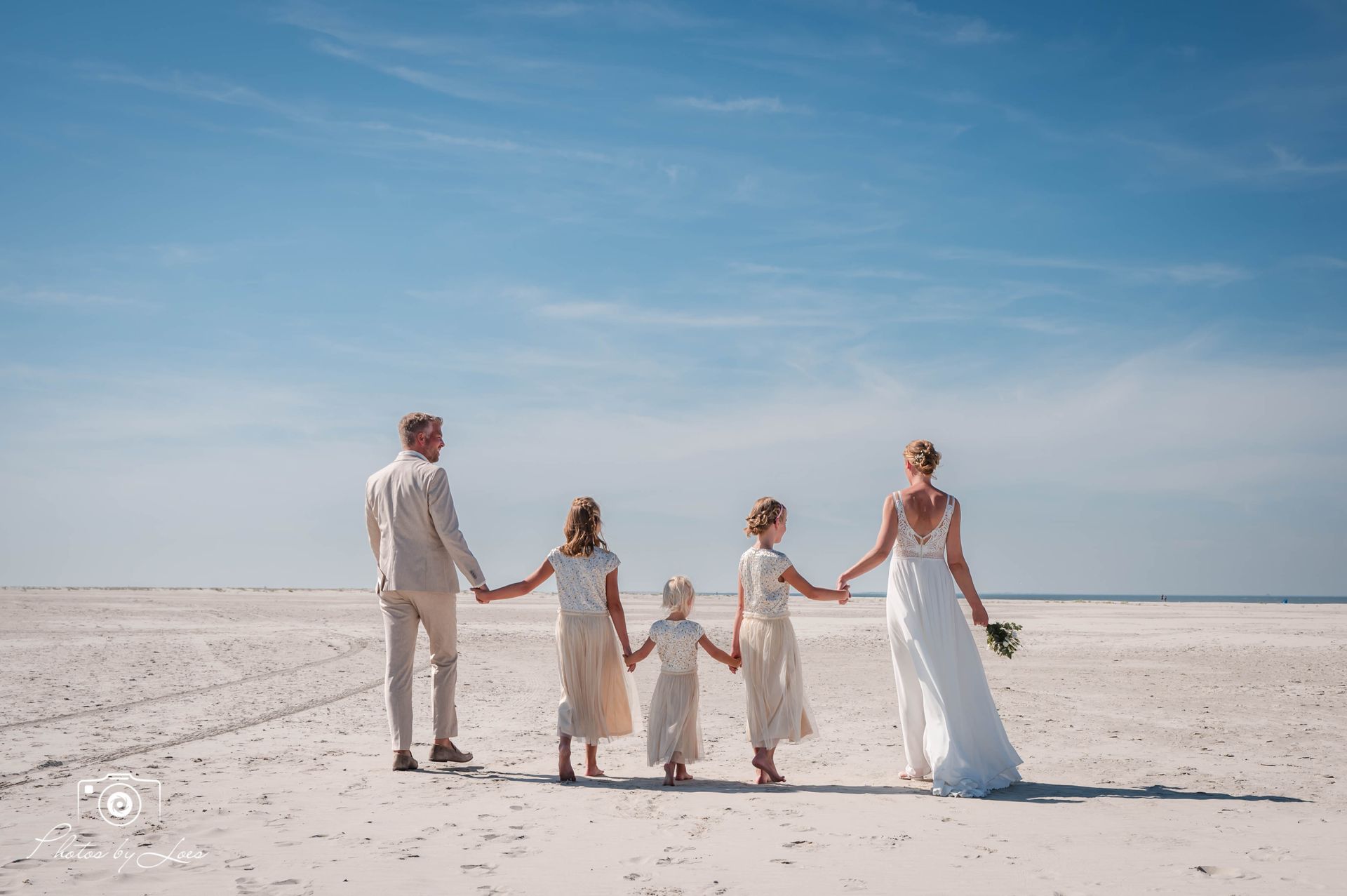Een bruid en bruidegom houden elkaars hand vast met hun bloemenmeisjes op het strand.