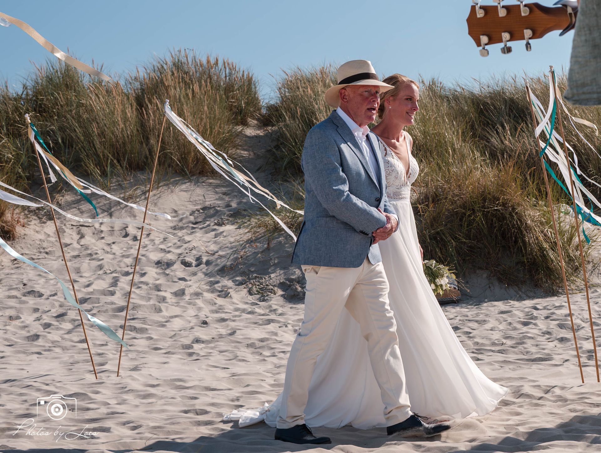 Een bruidspaar loopt over het strand met een gitaar op de achtergrond.