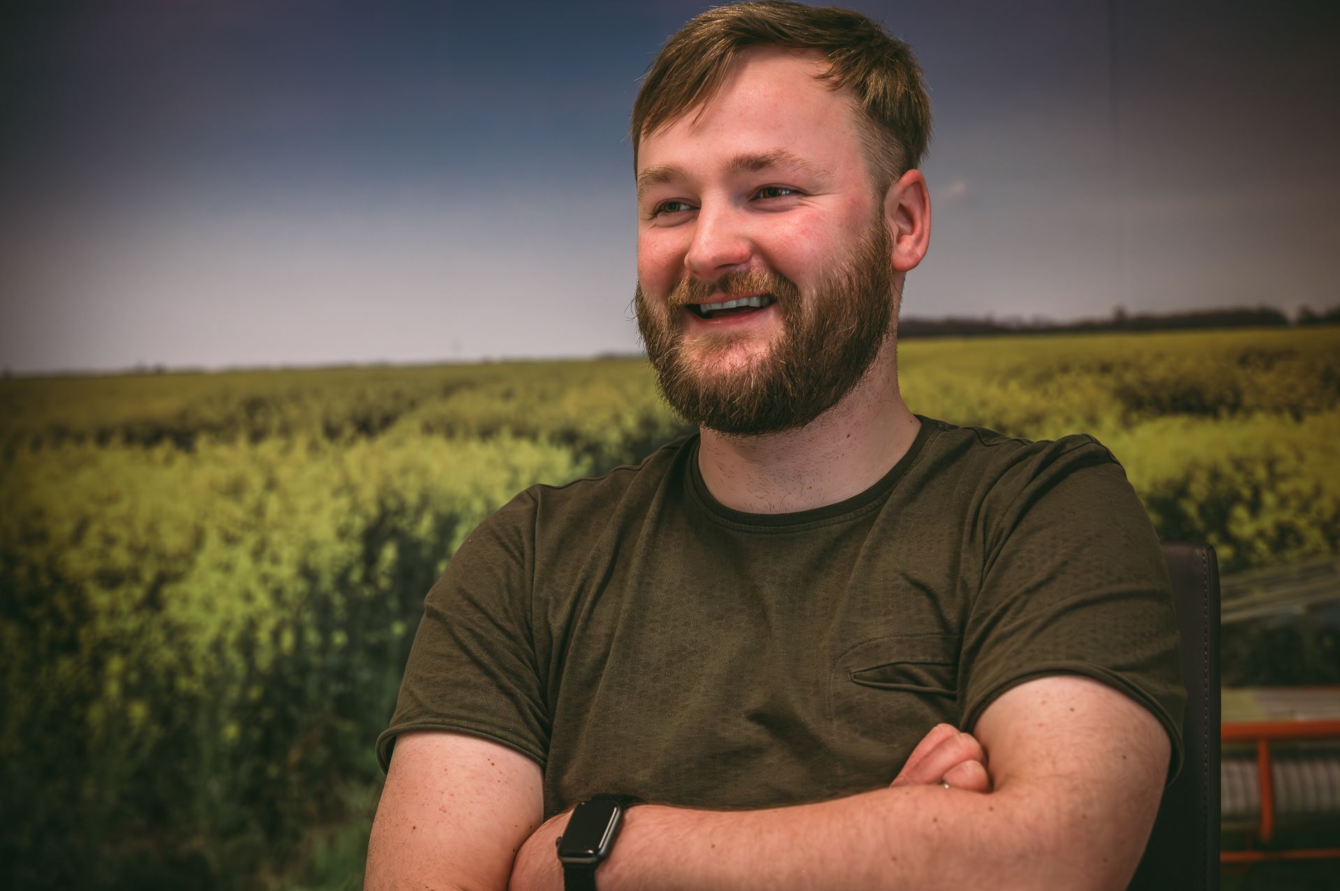 Een man met een baard staat lachend met zijn armen over elkaar voor een veld.