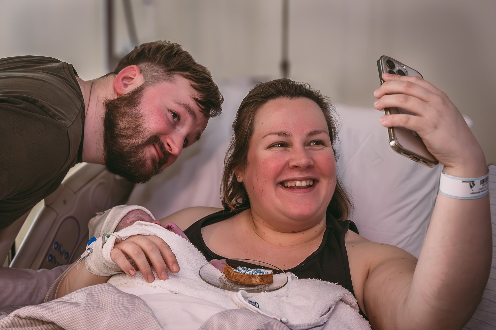 Een man en een vrouw maken een selfie met hun pasgeboren baby in een ziekenhuisbed.