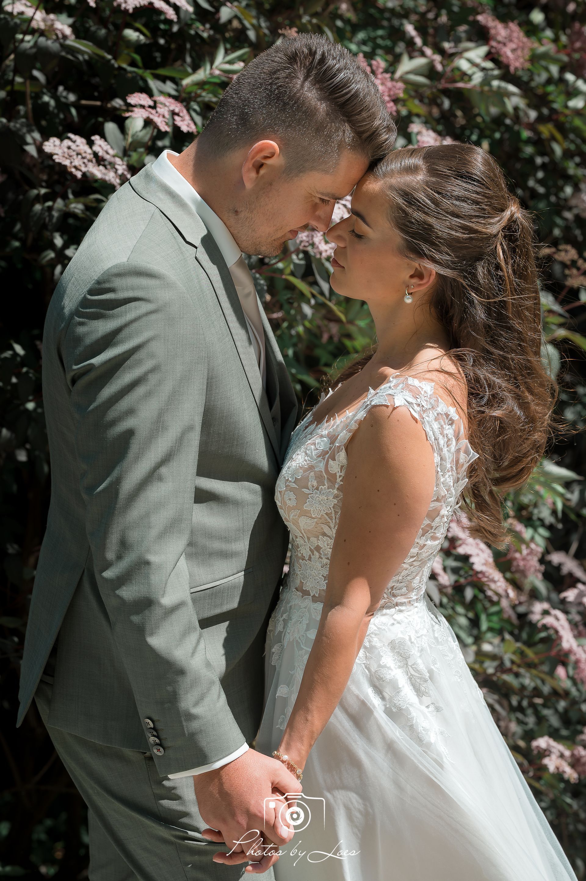 Een bruid en bruidegom kussen elkaar voor een struik met bloemen.