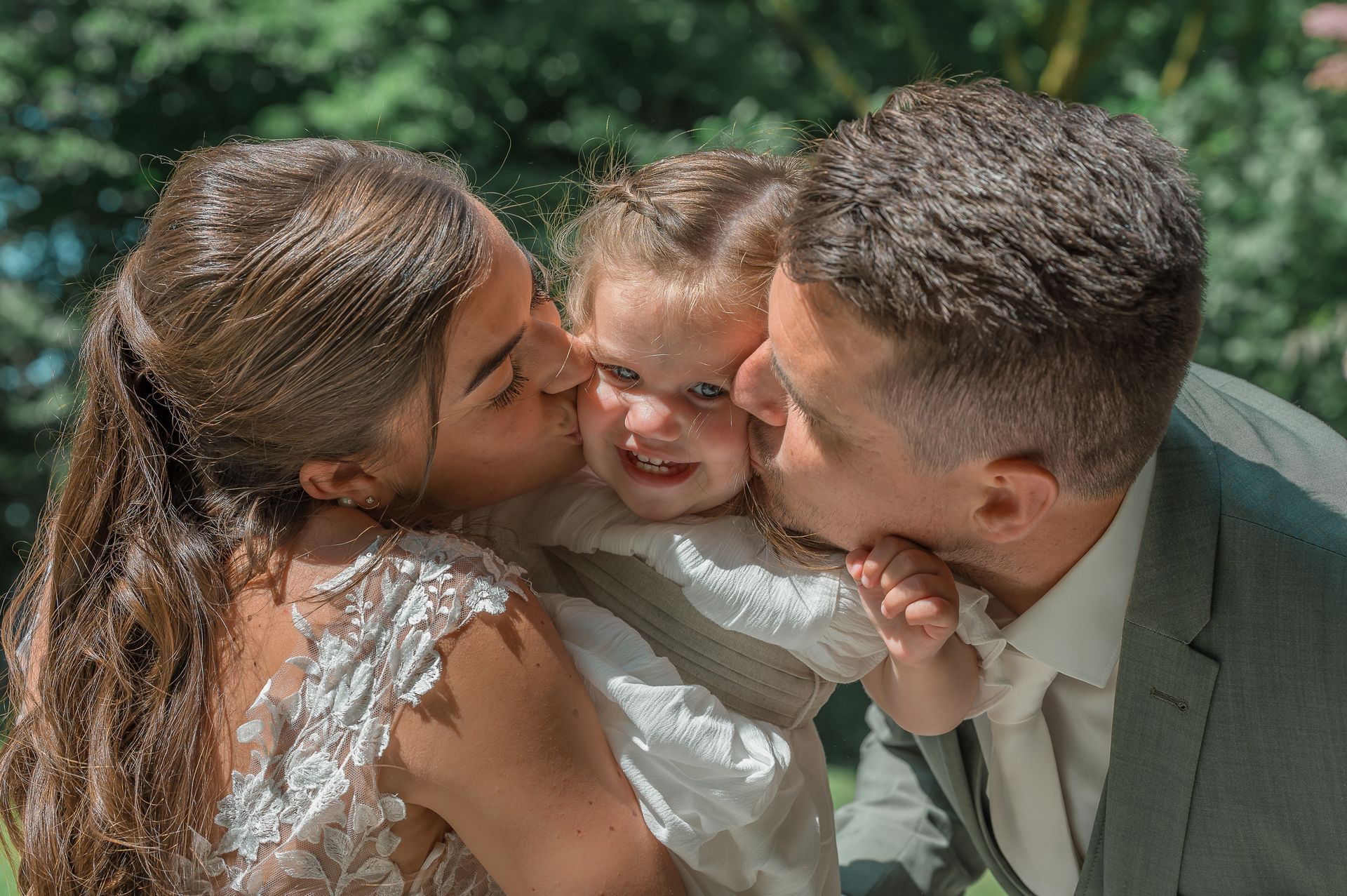 Een bruid en bruidegom kussen hun dochter op de wang.