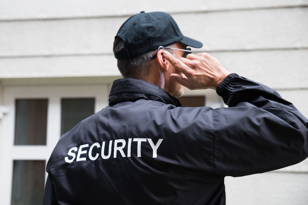 Guardia de seguridad con chaqueta negra y gorra, escuchando por auricular y mirando hacia otro lado de la cámara.