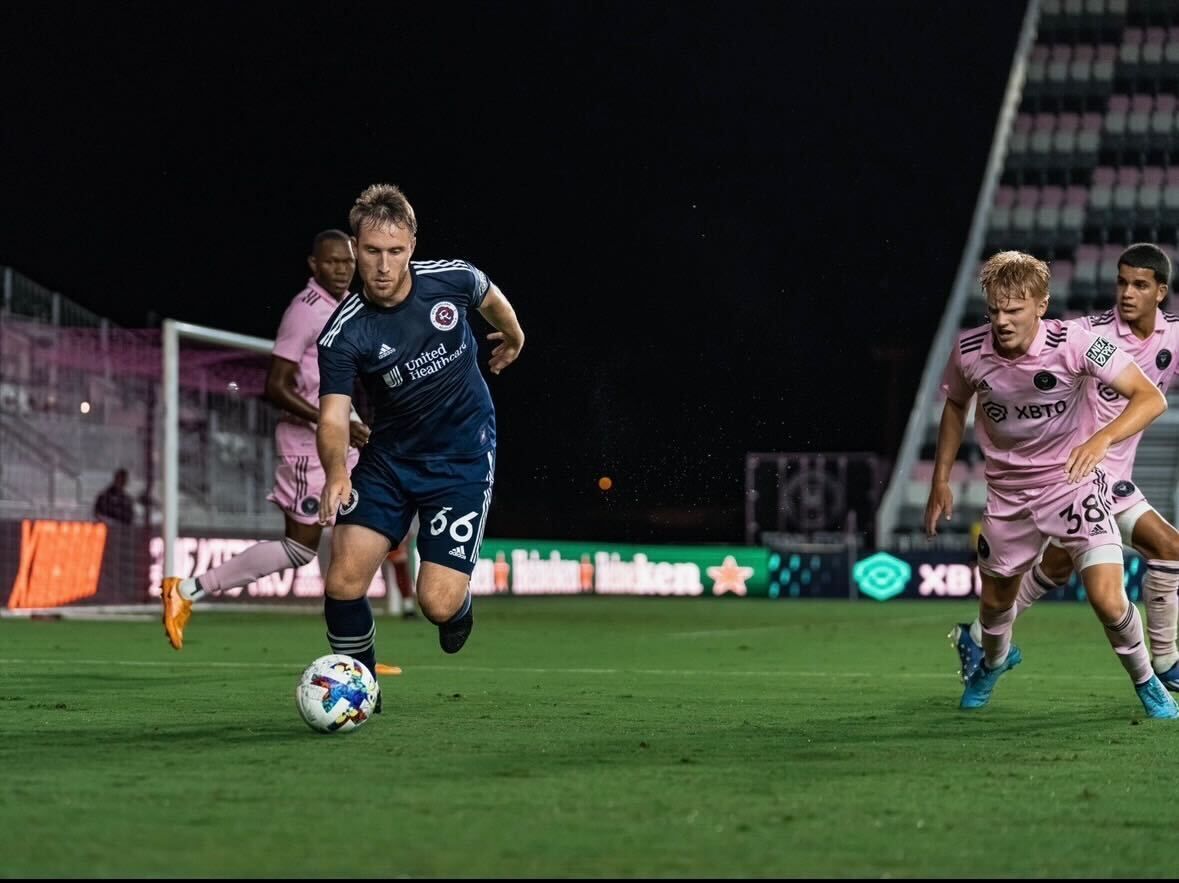 A player in a blue kit dribbles a soccer ball at night while being pursued by opponents in pink jerseys on a stadium field.