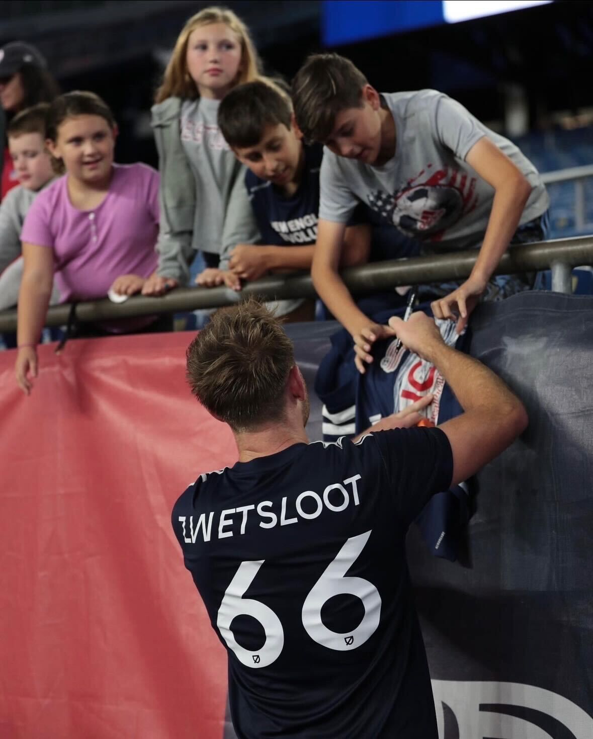 A jersey-wearing athlete signs an autograph for a group of fans leaning over a stadium railing.