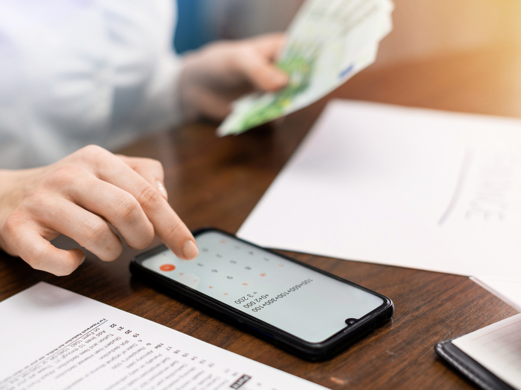 Person counting money and using a calculator app on a smartphone on a wooden table.